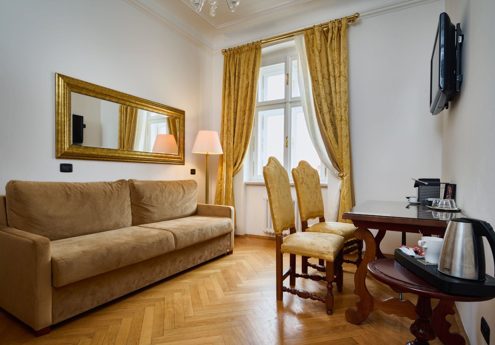 A hotel room with a couch, table, chairs, window, and TV. The walls are white with gold curtains.