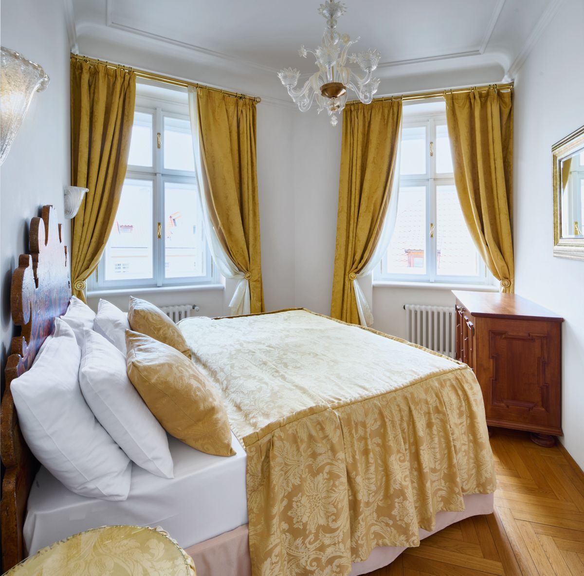 Bedroom with gold curtains, bedspread, and wooden furniture. Two windows and ornate chandelier.