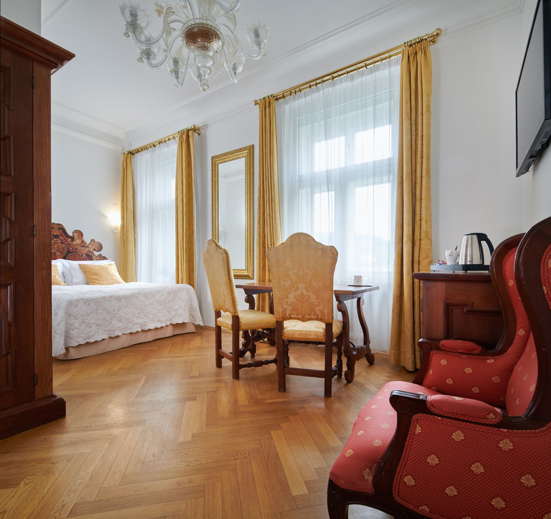Hotel room with ornate furniture, including a bed, table, chairs, and armoire. Gold curtains and a chandelier are visible.