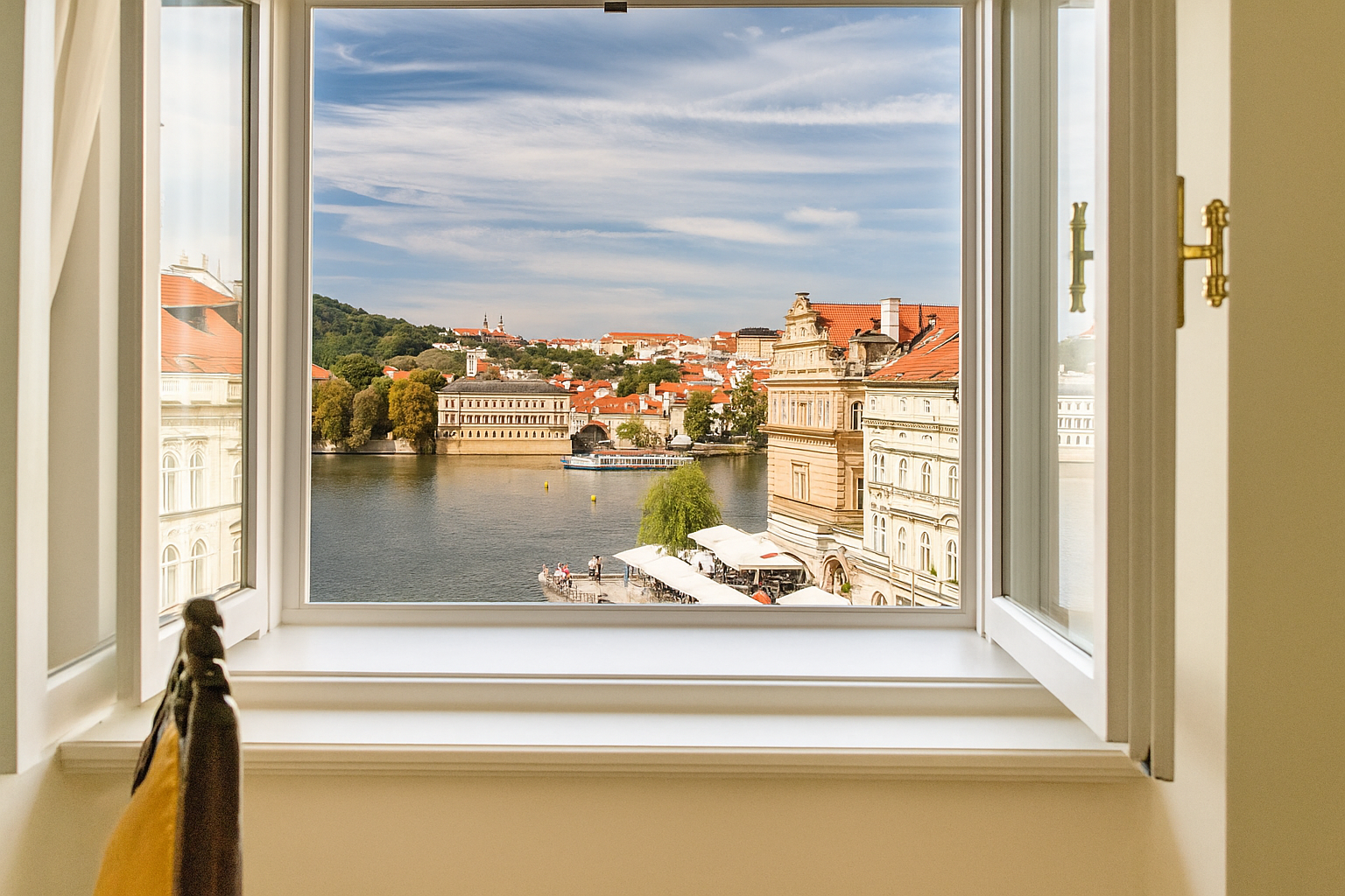 Open window framing a view of a European cityscape with a river, buildings, and blue sky.