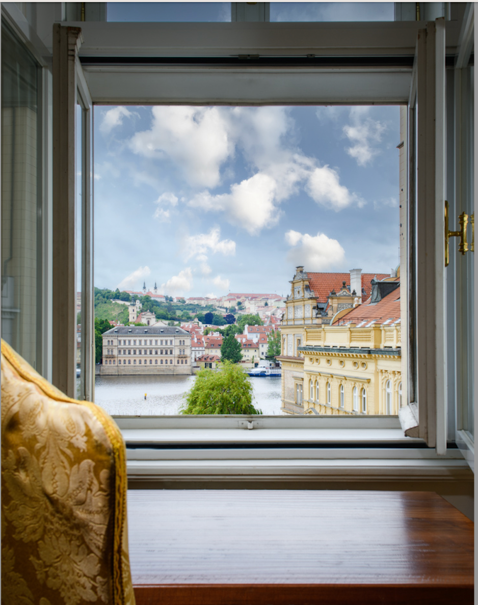 View of Prague from an open window, overlooking the Vltava River and city buildings, under a blue sky.