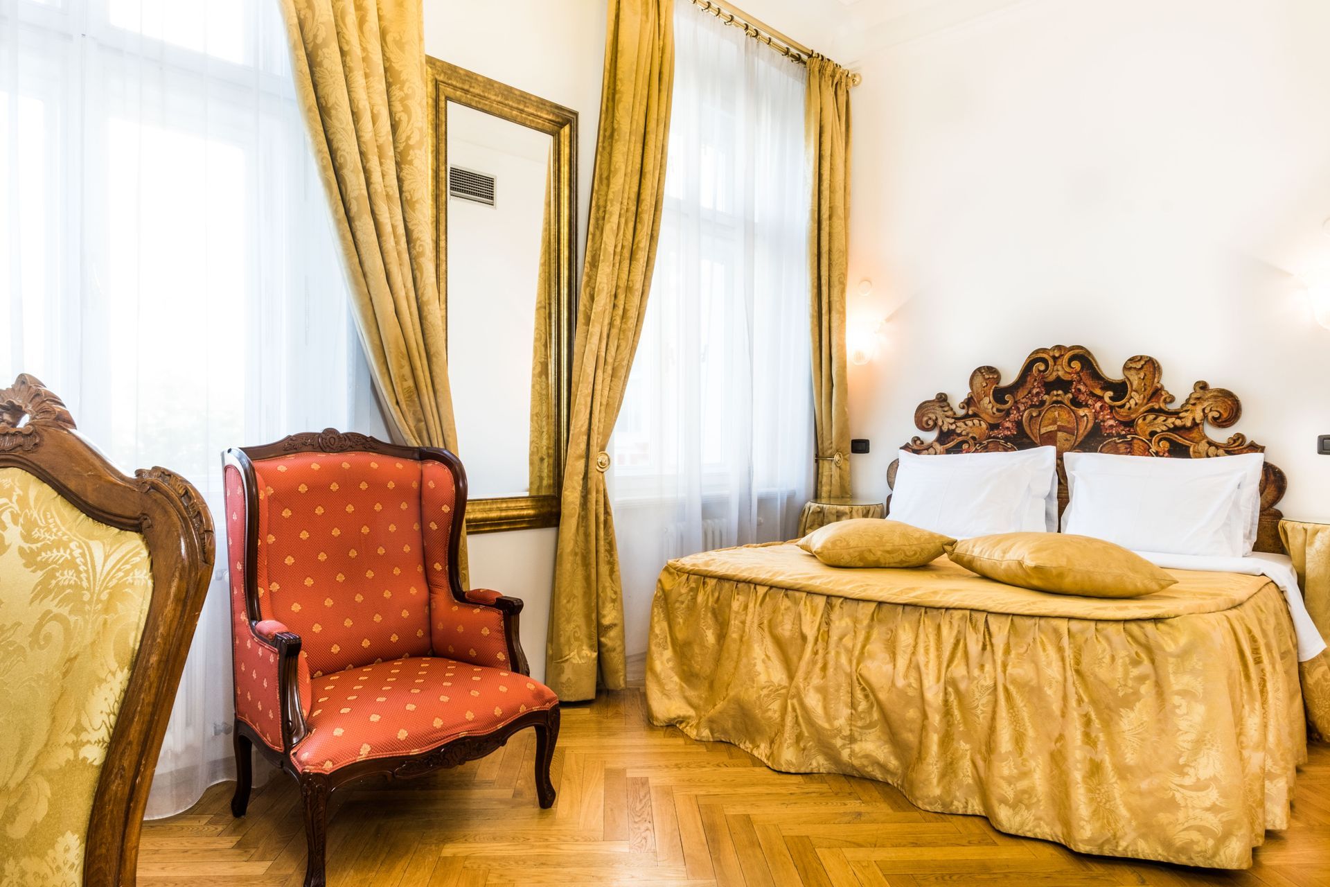 Elegant hotel room with ornate bed, gold accents, red armchair, and large mirror.
