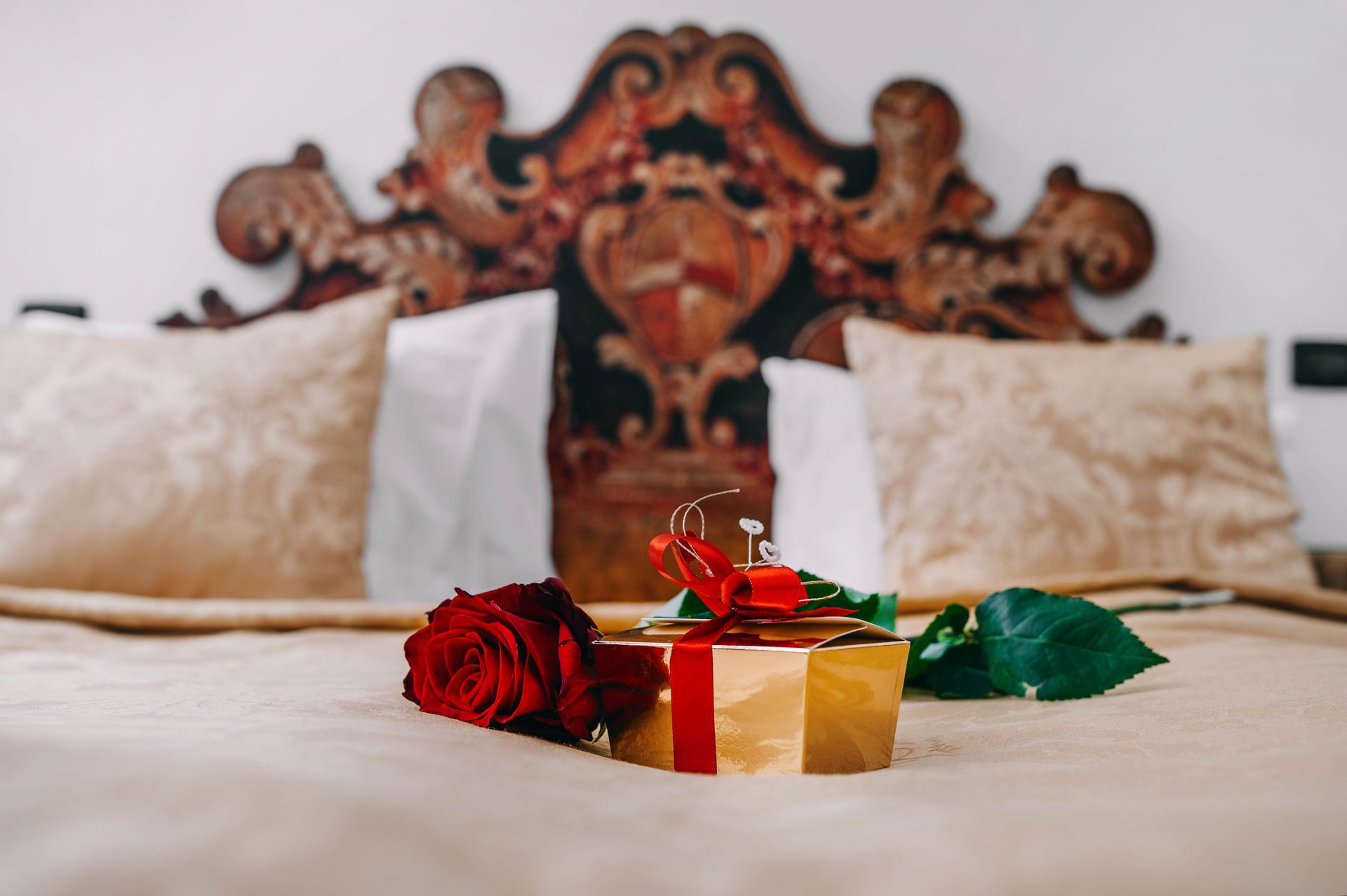 Red rose, wrapped gift, and ring on a bed with ornate headboard.