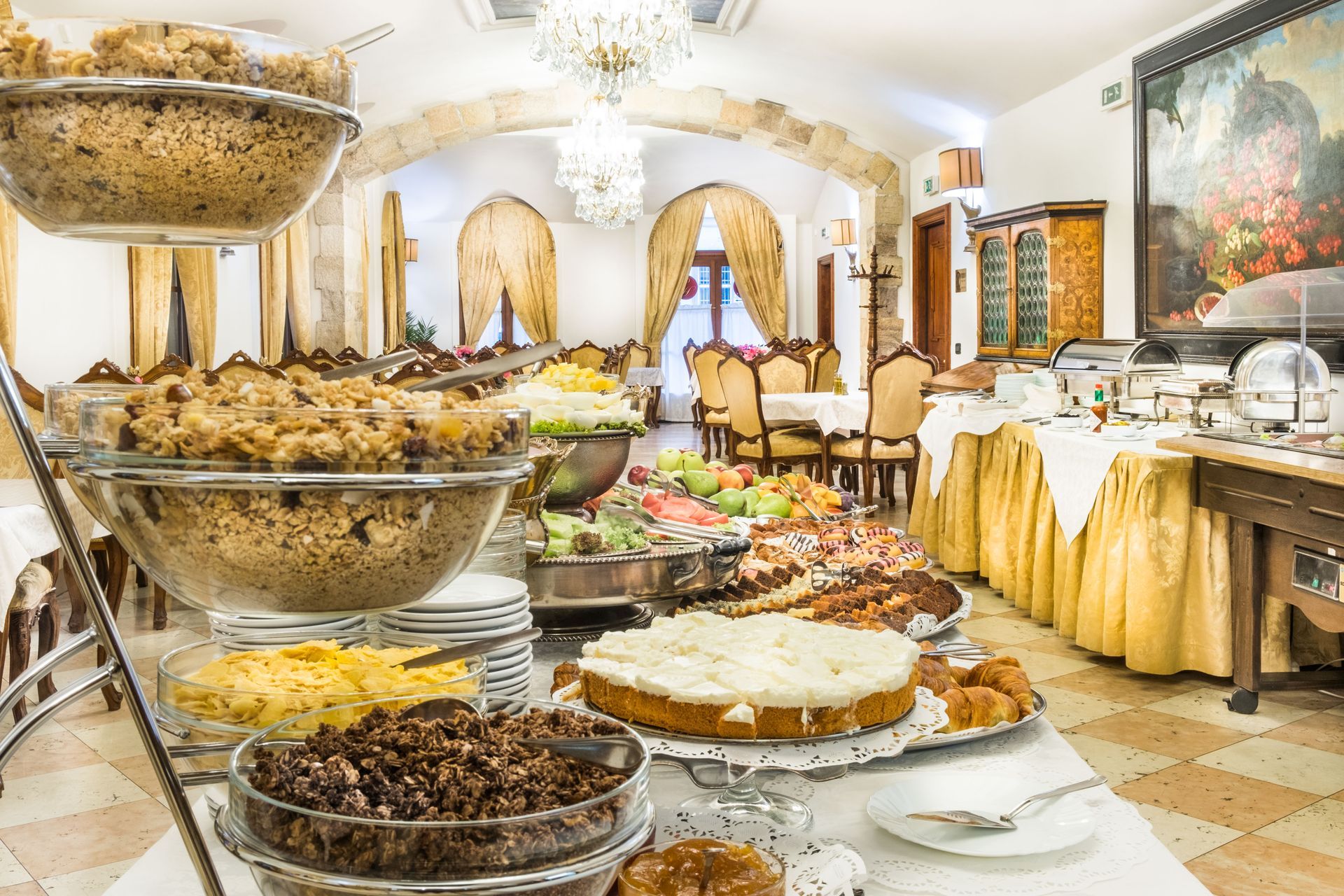 Colazione a buffet con dolci, frutta e piatti caldi in una sala da pranzo impreziosita da lampadari e opere d'arte.