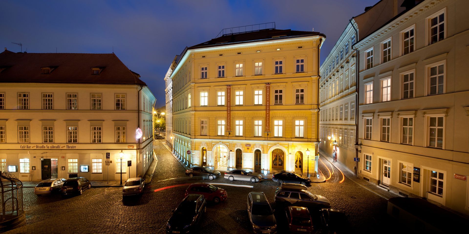 Cobblestone street at night, lined with illuminated buildings, cars parked, and a central building with arched entry.