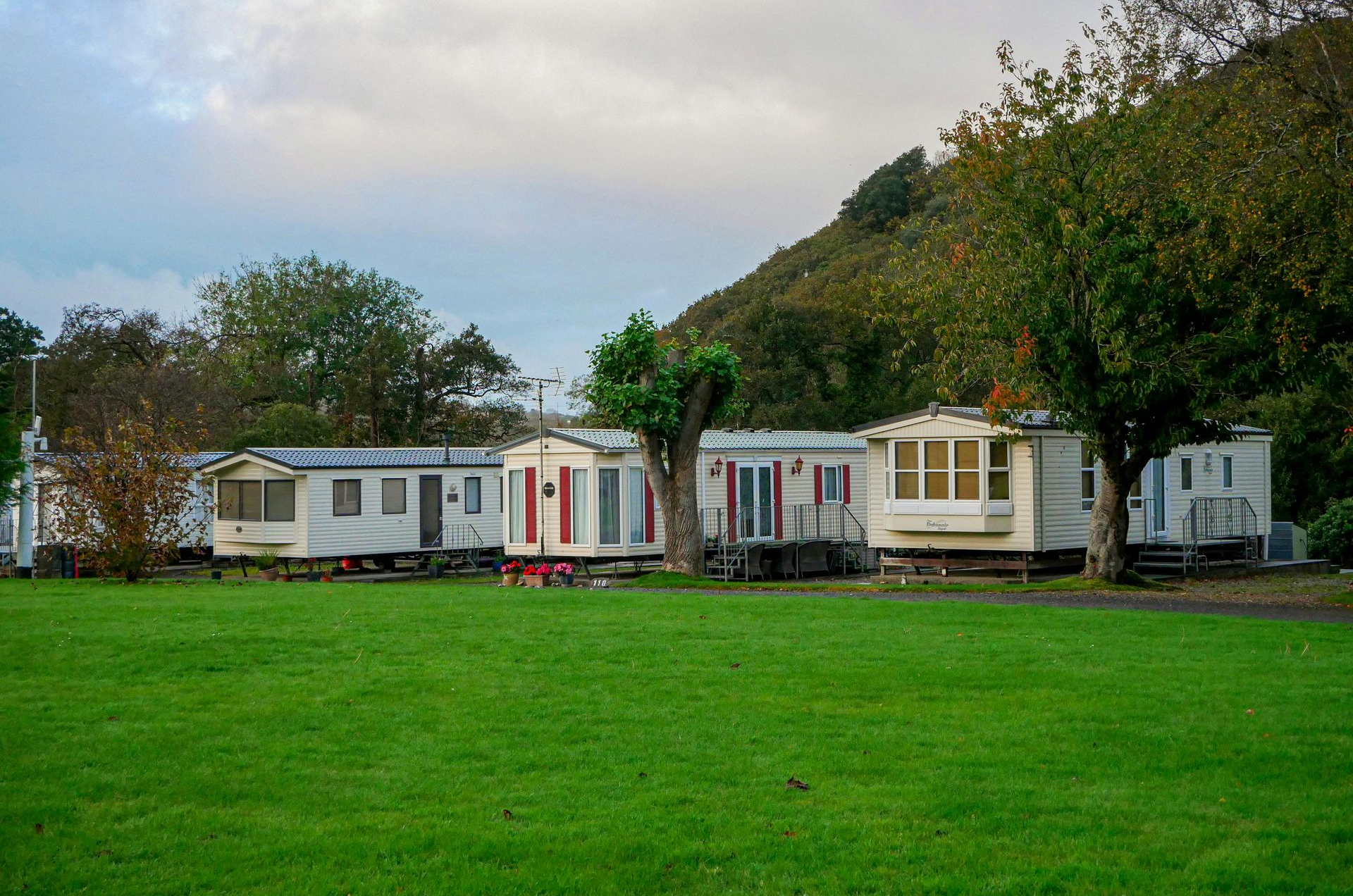 Row of white caravans beside a large green lawn and trees under a cloudy sky