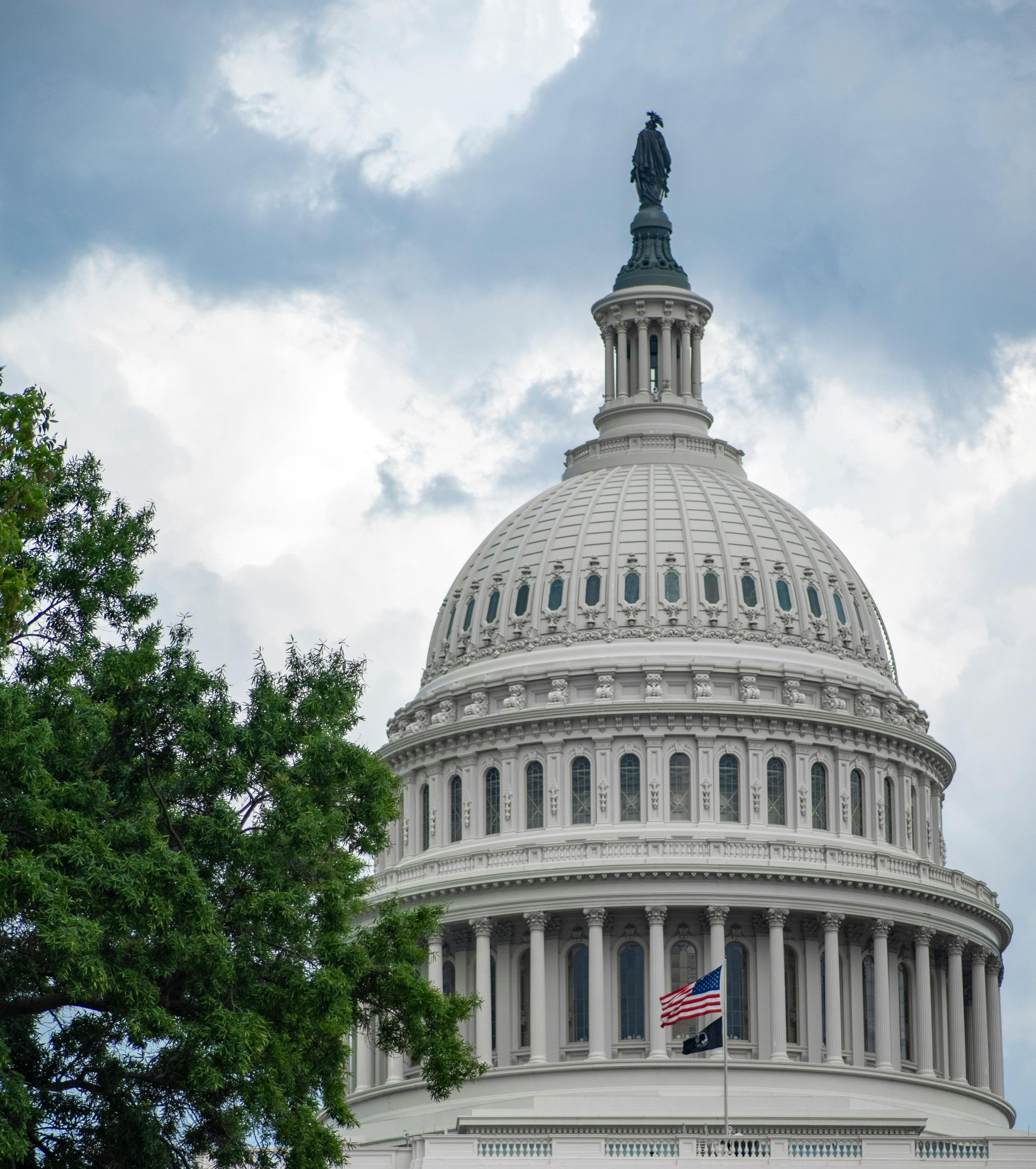 U.S. Capitol dome against a cloudy sky, framed by trees, with an American flag below