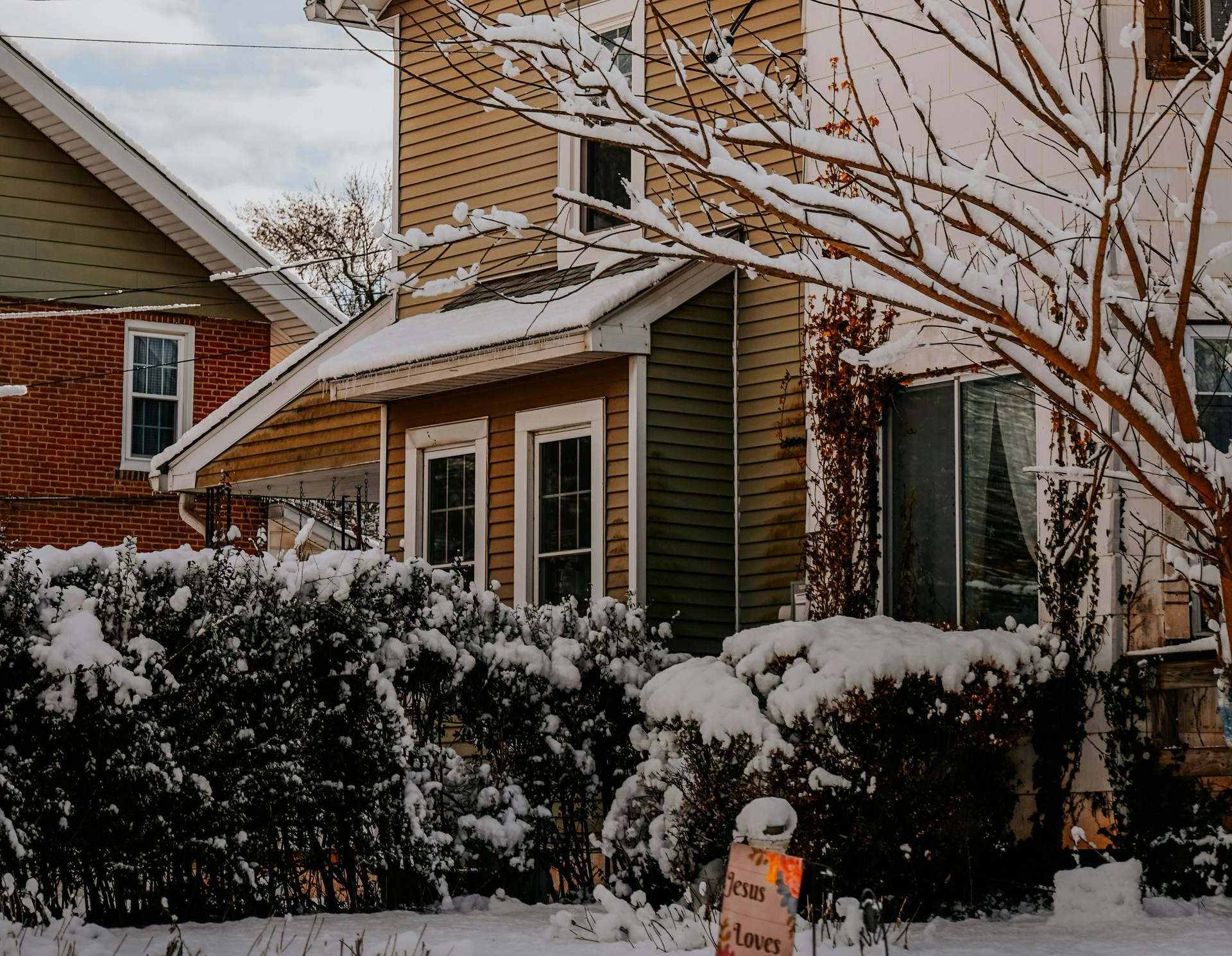 Snow-covered suburban house and bushes with a small yard sign in front.
