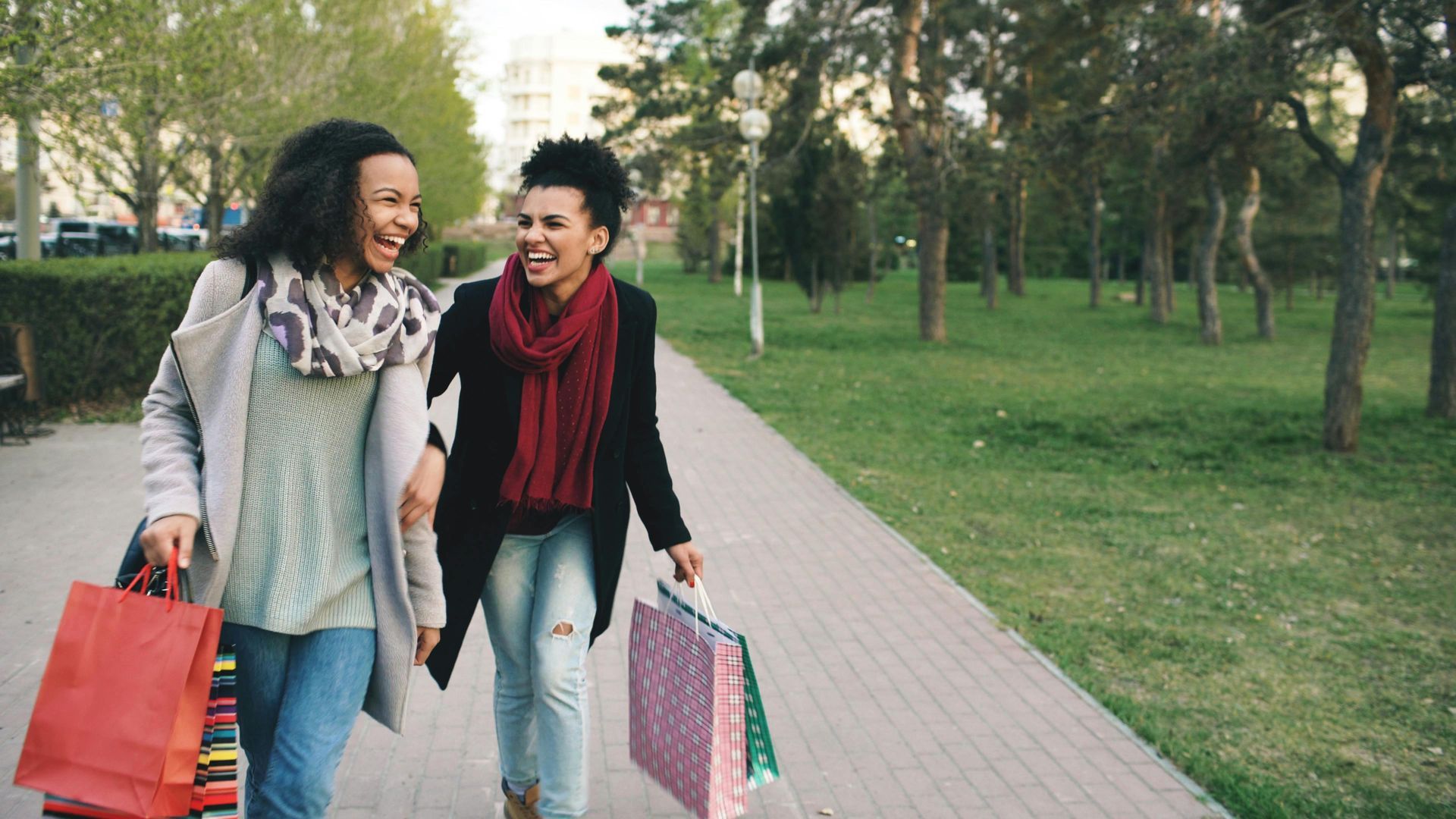 Two friends walking and smiling on a park path, carrying colorful shopping bags.