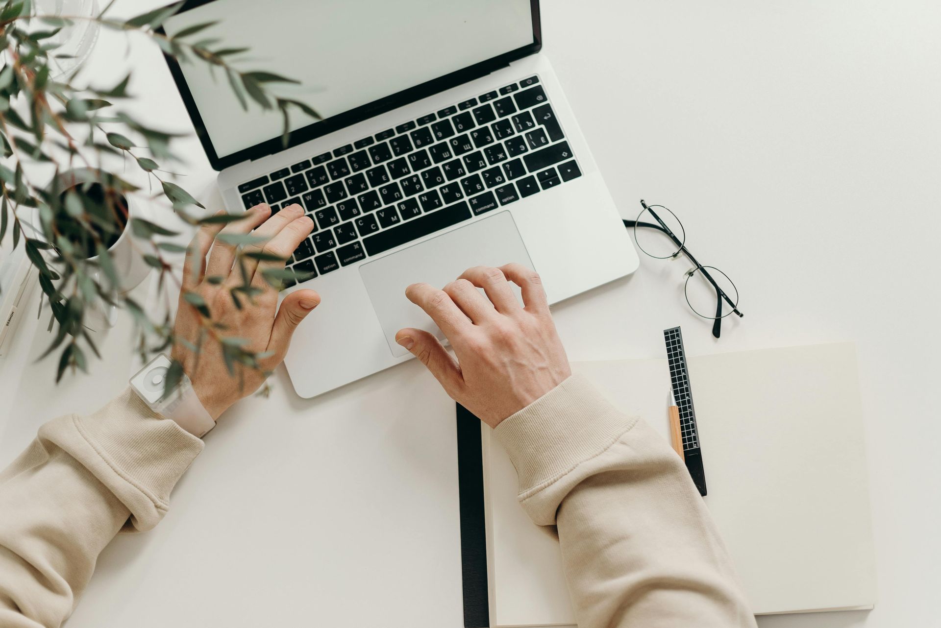 Hands typing on a laptop beside an open notebook on a white desk with a plant.
