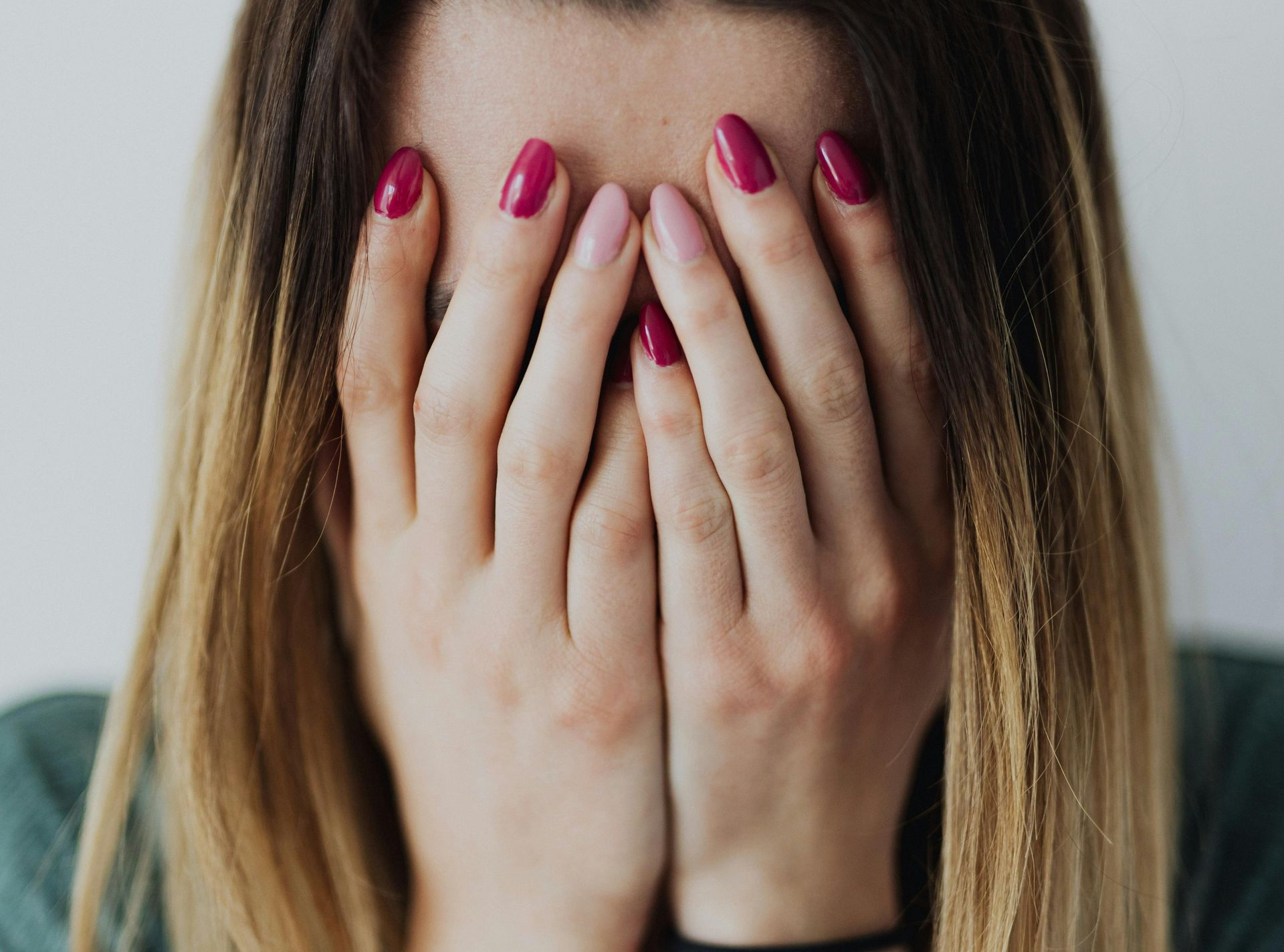 Person covering their face with both hands, wearing pink nail polish, against a blurred background.
