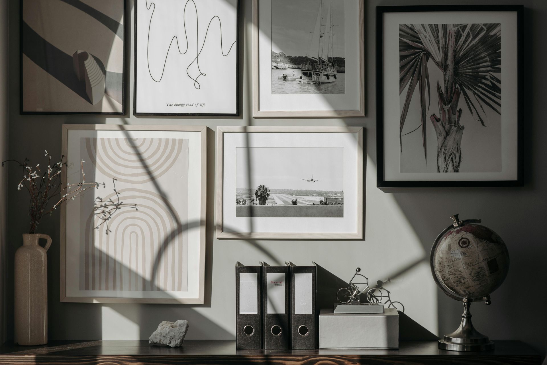 Framed black-and-white art on a sunlit wall above a shelf with books, plants, and a globe.
