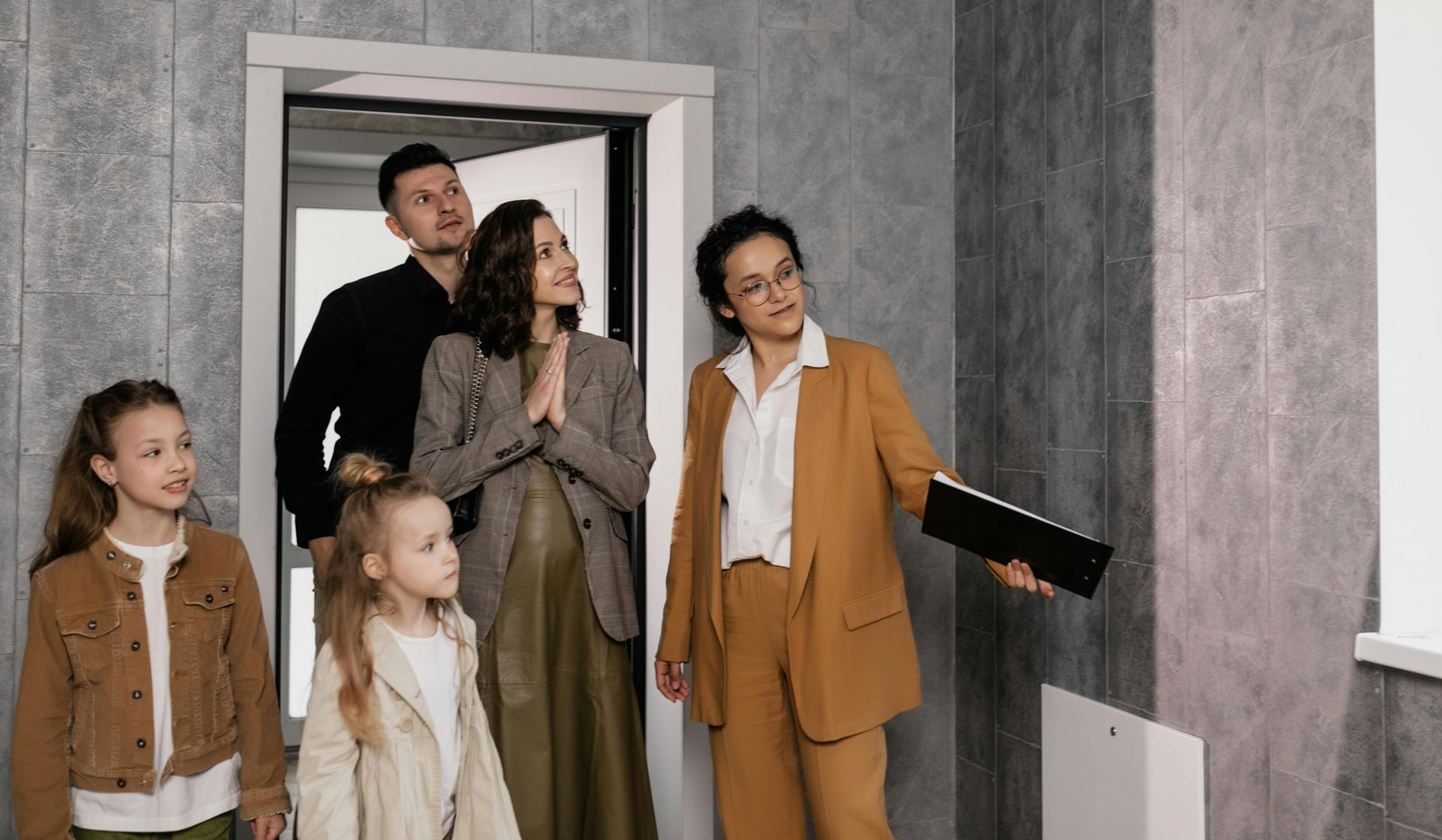 Four people stand in a doorway, looking into a gray-walled room; one holds a black folder.