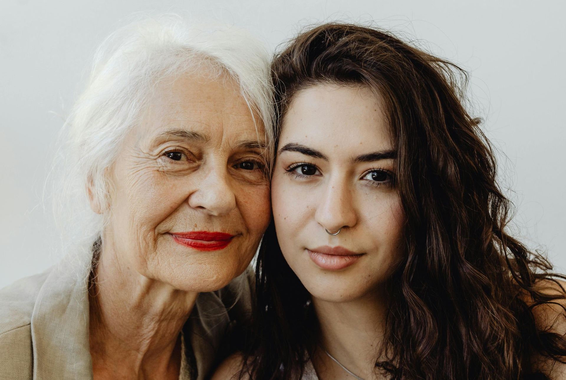 Two women pose closely, one with white hair and red lipstick, in a softly lit portrait.