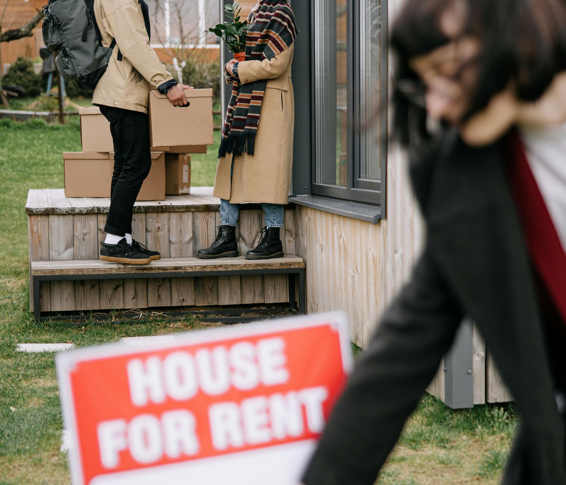 Two people talking on a porch beside a house for rent sign.