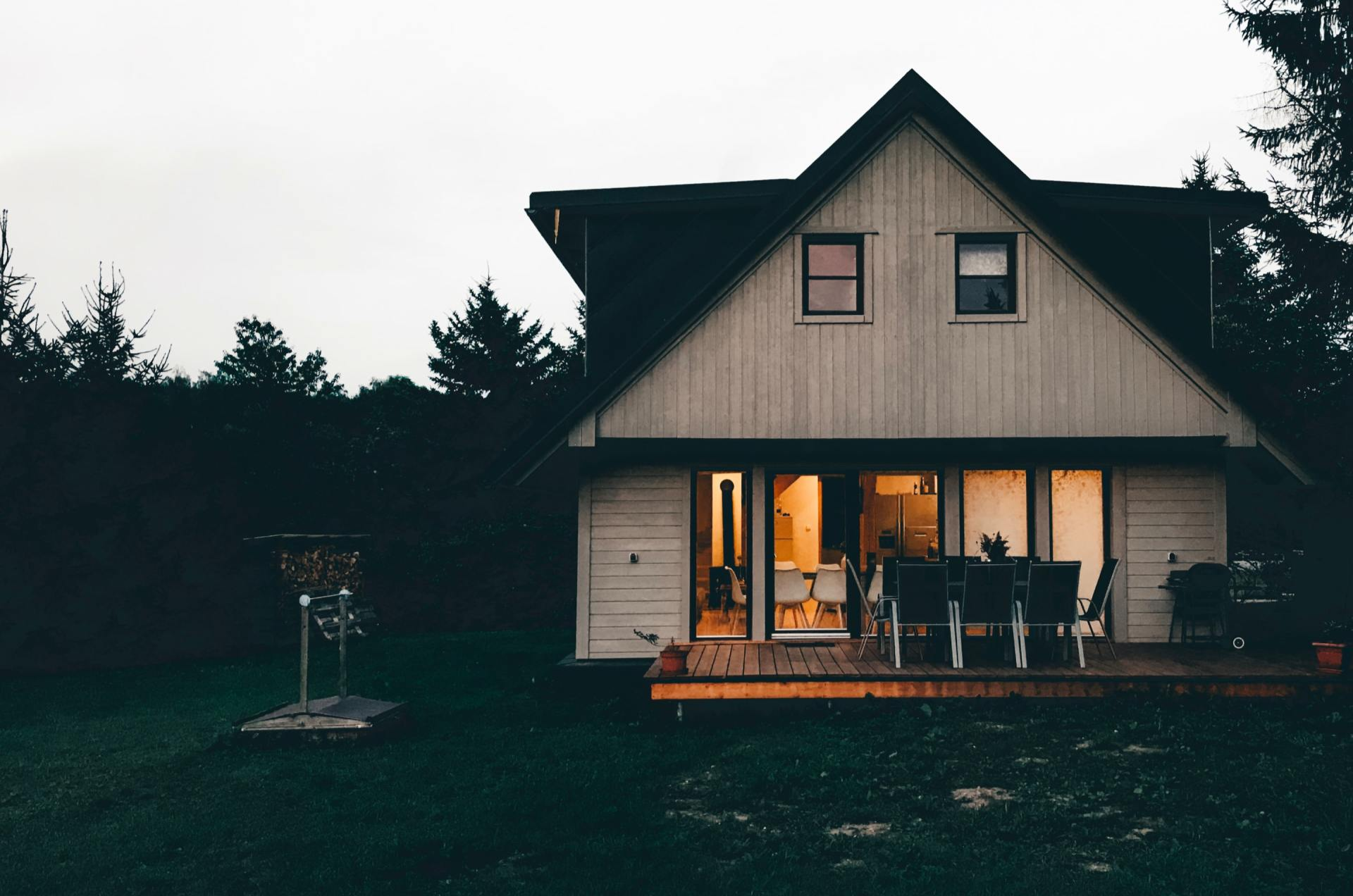 Cabin-style house at dusk with lit porch and outdoor seating, surrounded by trees