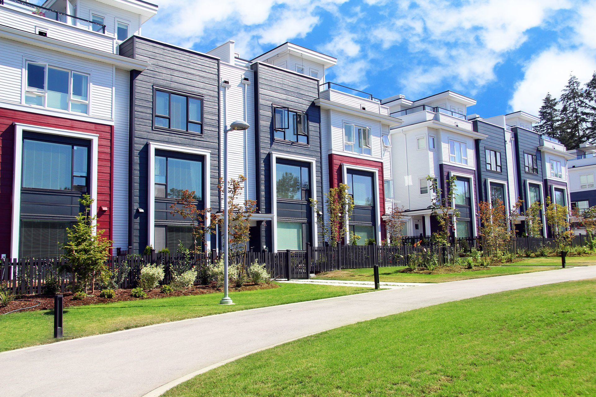 Modern townhomes with gray, red, and white facades beside a sidewalk and green lawn under a blue sky