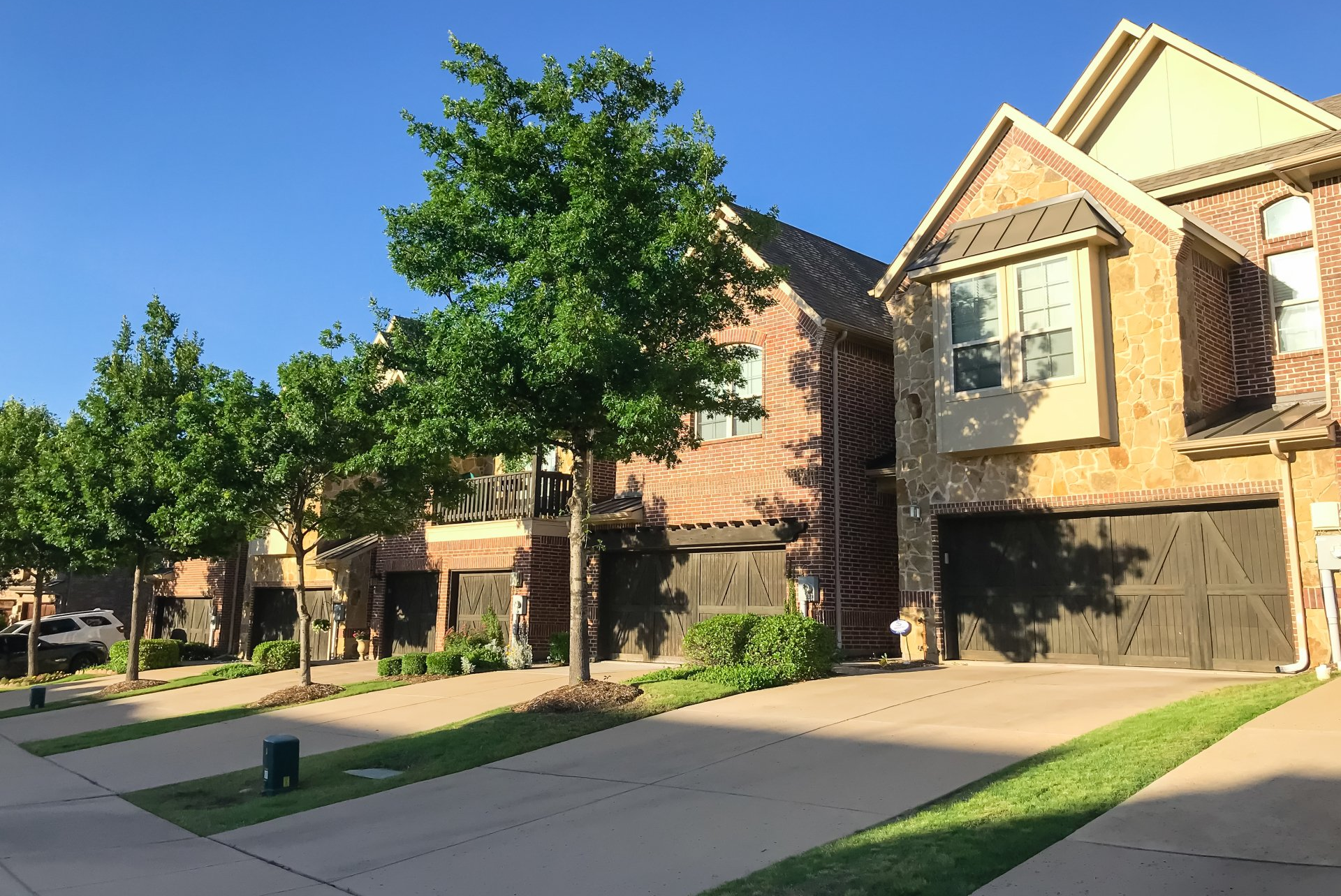 Sunlit suburban street with brick townhouses, driveways, and trees along the sidewalk