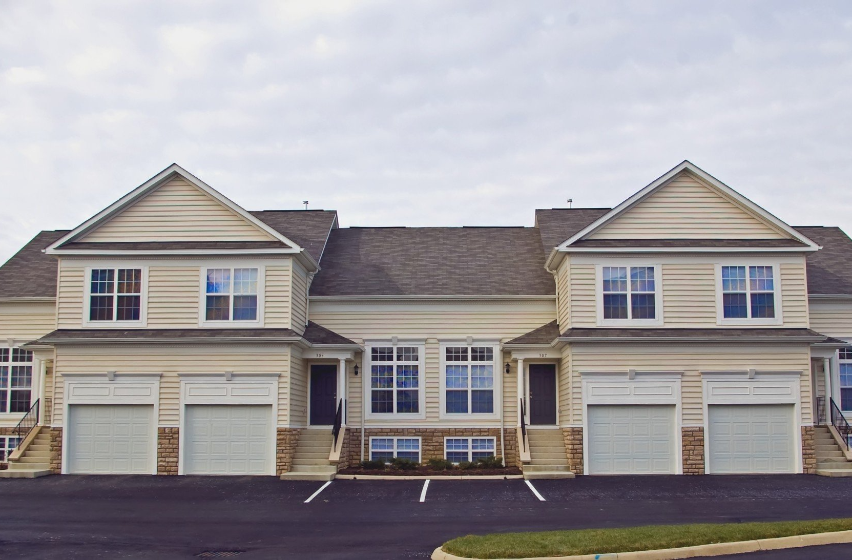 Two beige apartment buildings with garages and a central entrance, viewed from a parking lot.