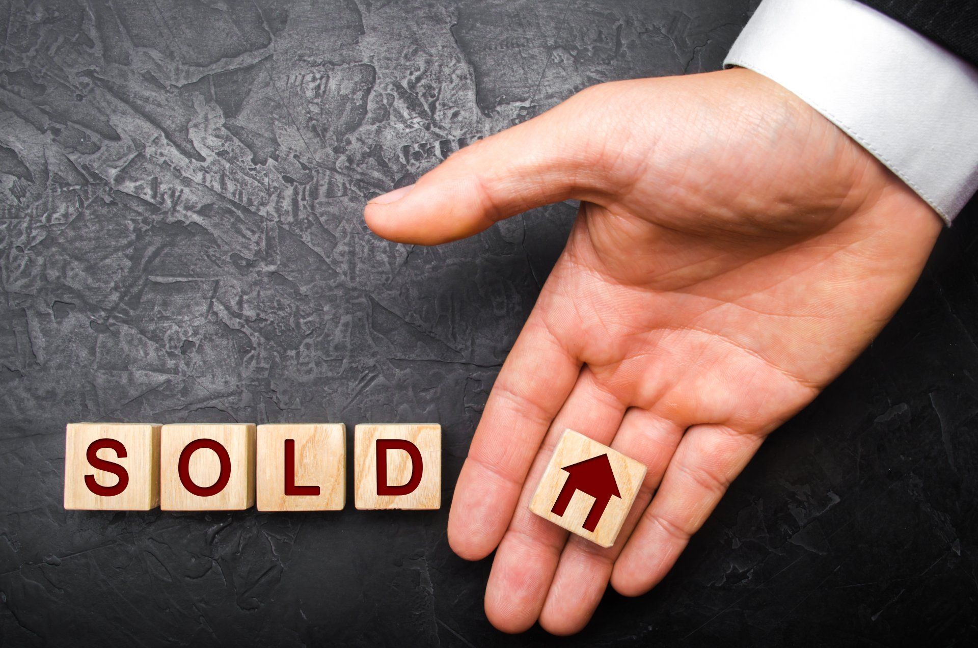 Hand holding a small house token beside letter blocks spelling “SOLD” on a dark background