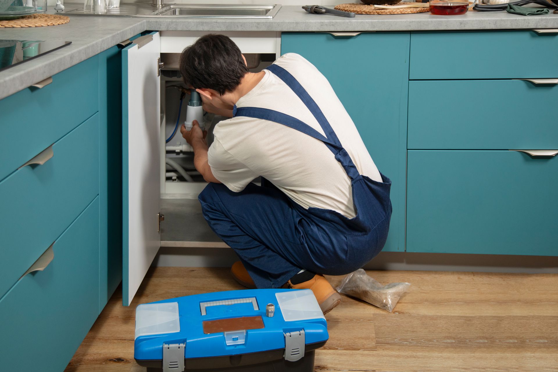 A person in blue overalls kneels fixing an appliance in a kitchen with blue cabinets. A toolbox sits nearby.