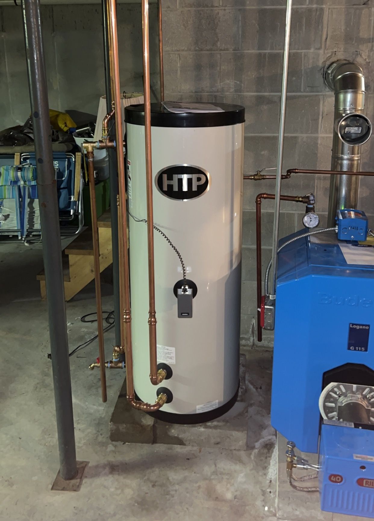 A htp water heater is sitting in a basement next to a blue boiler.