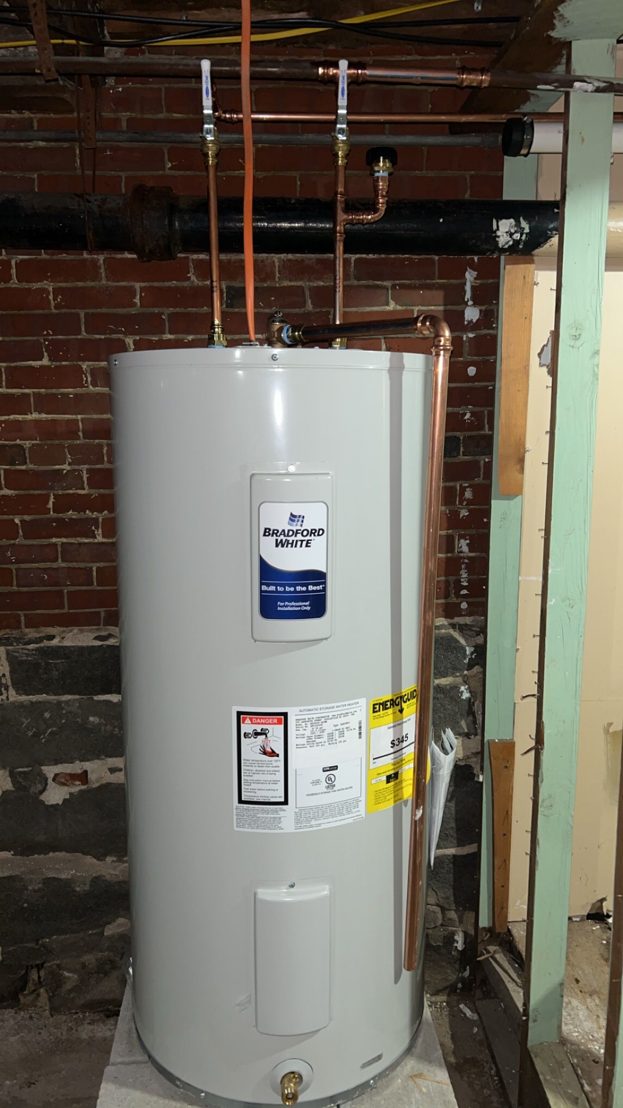 A water heater is sitting in a basement next to a brick wall.