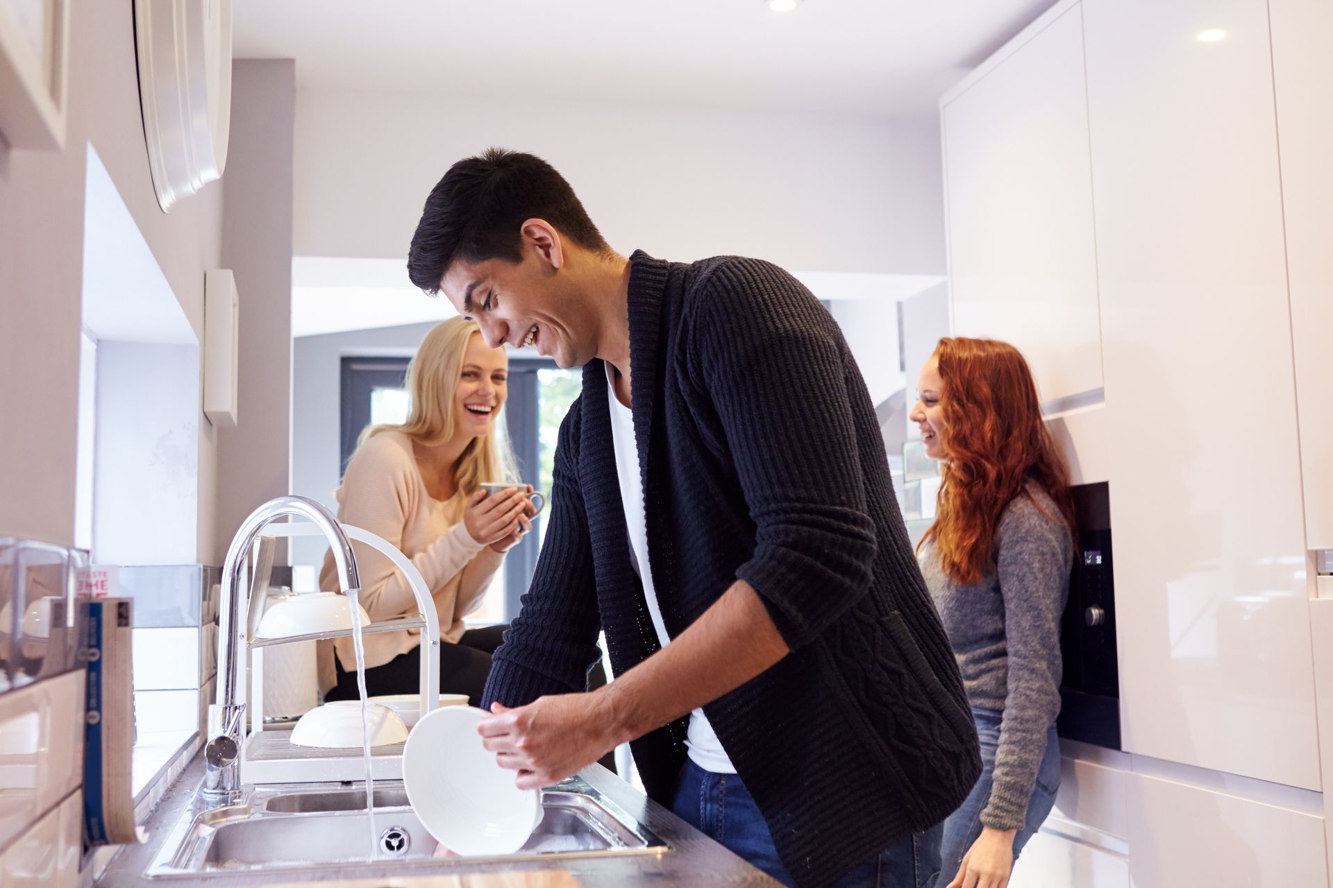 A man is washing dishes in a kitchen while two women look on.