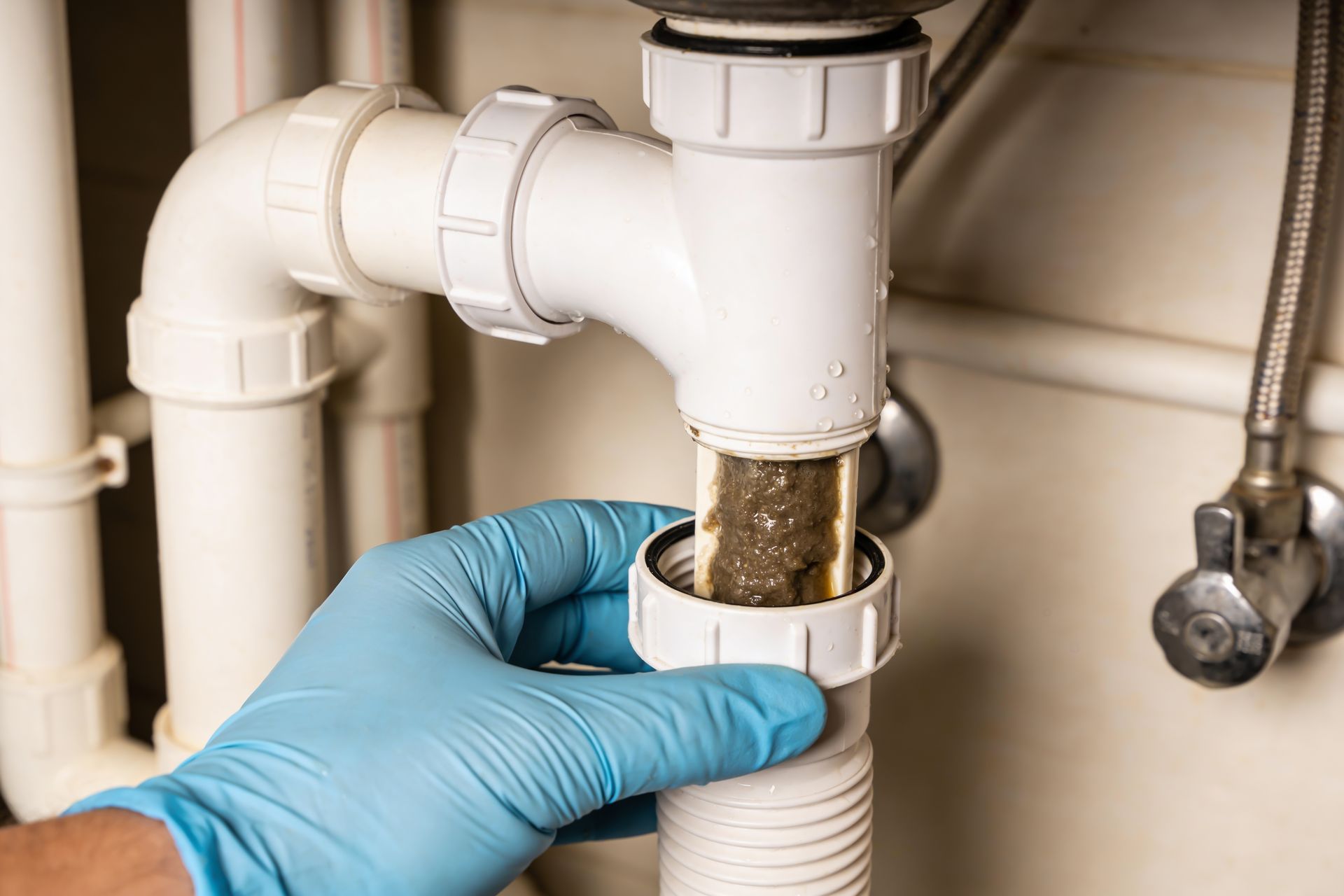 Gloved hands inspecting a clogged white sink drain under a bathroom sink