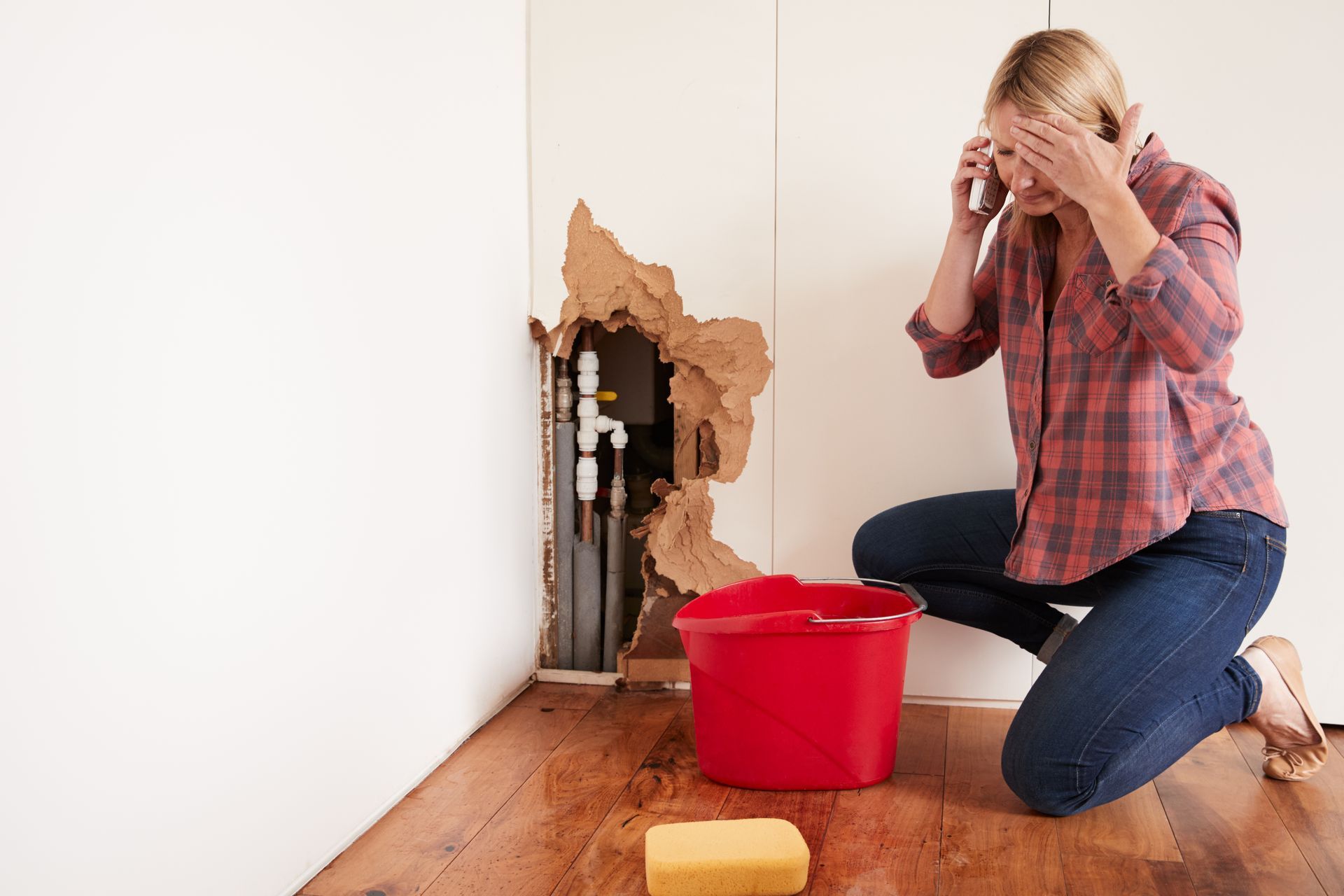 Person kneeling by a damaged wall with water leak, holding head beside red bucket and sponge on floor