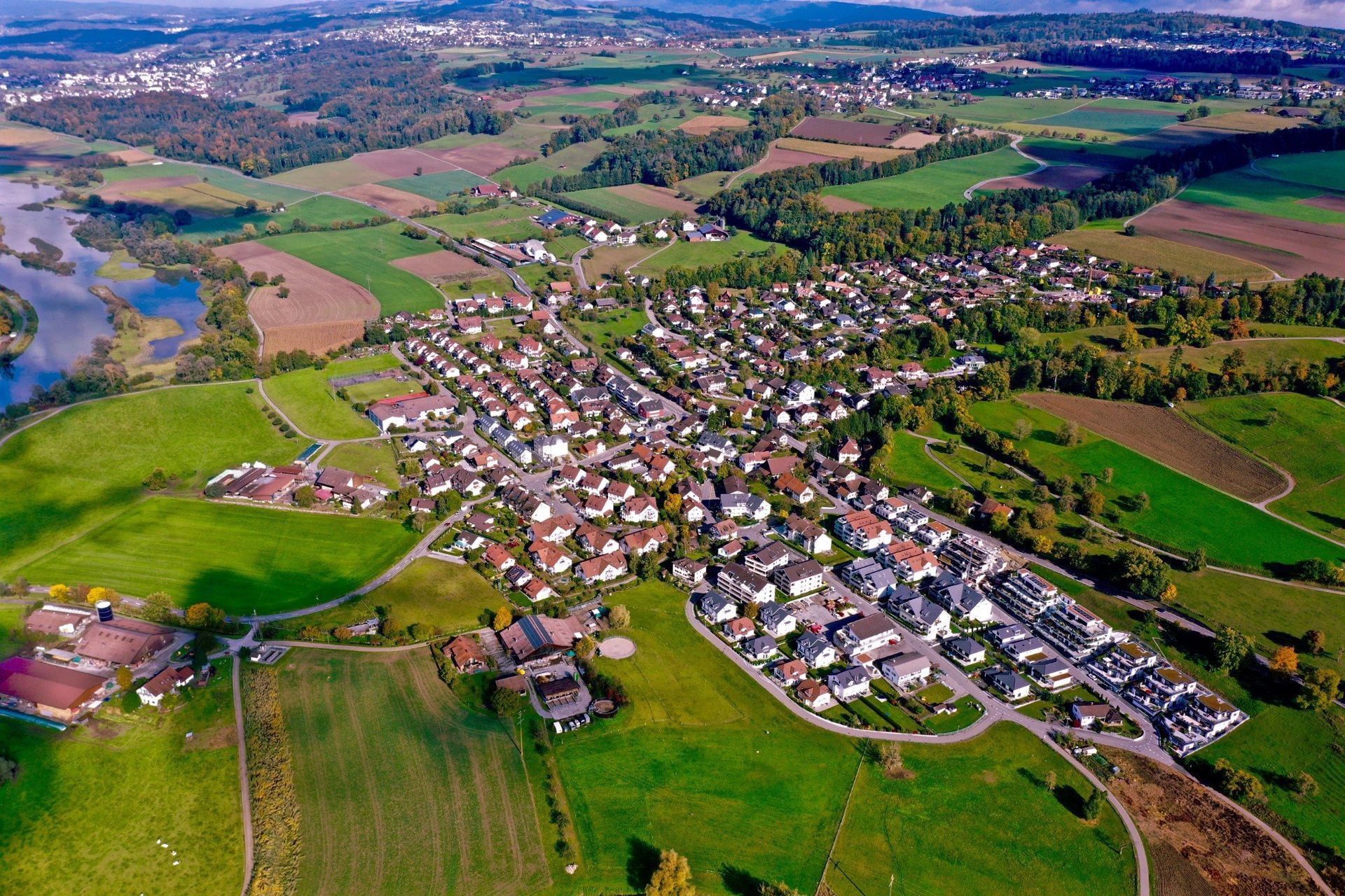 Luftaufnahme einer Stadt, eingebettet in grüne Felder, mit einem See und bewaldeten Hügeln in der Ferne.