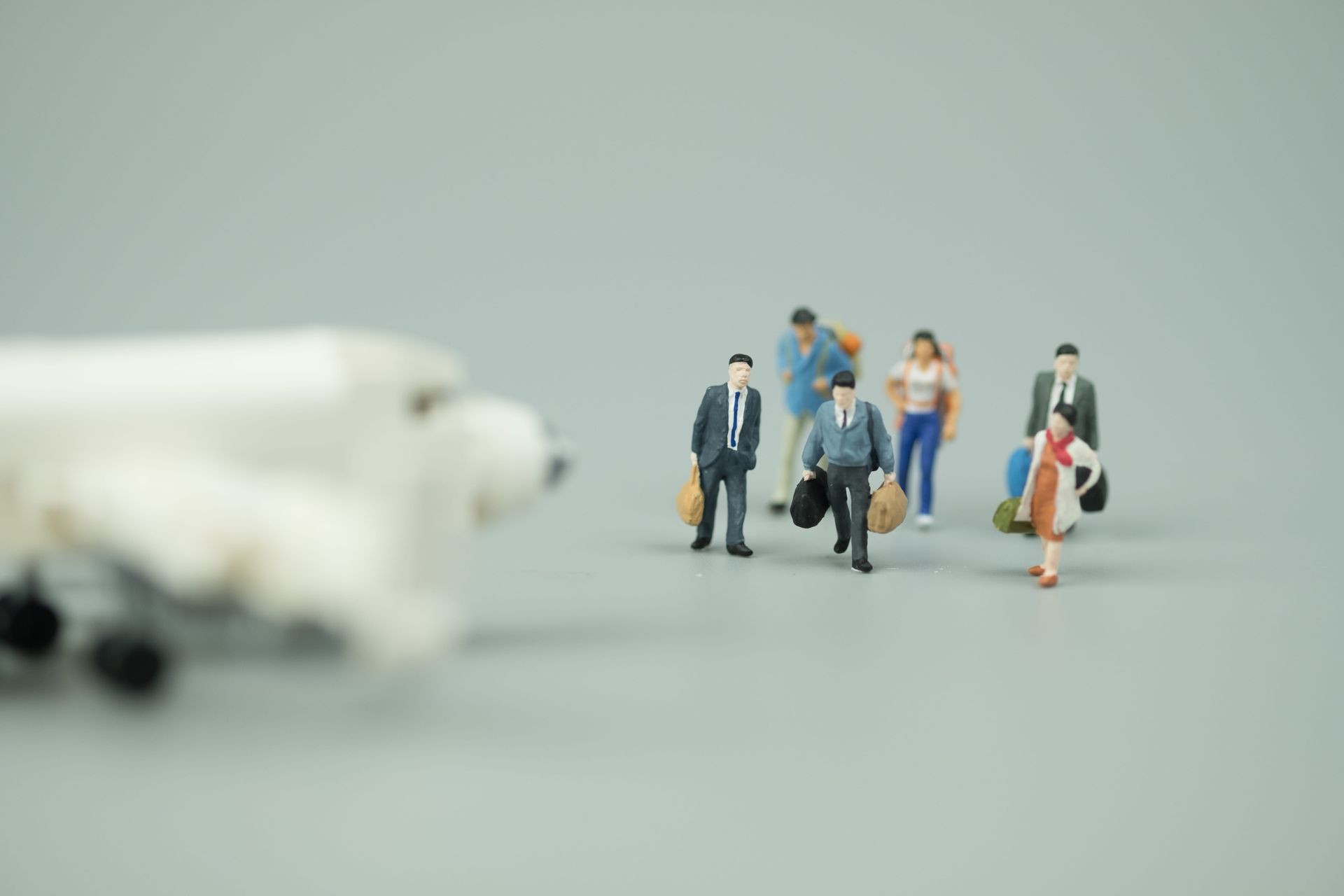 Miniature travelers with luggage beside a blurred airplane on a gray background