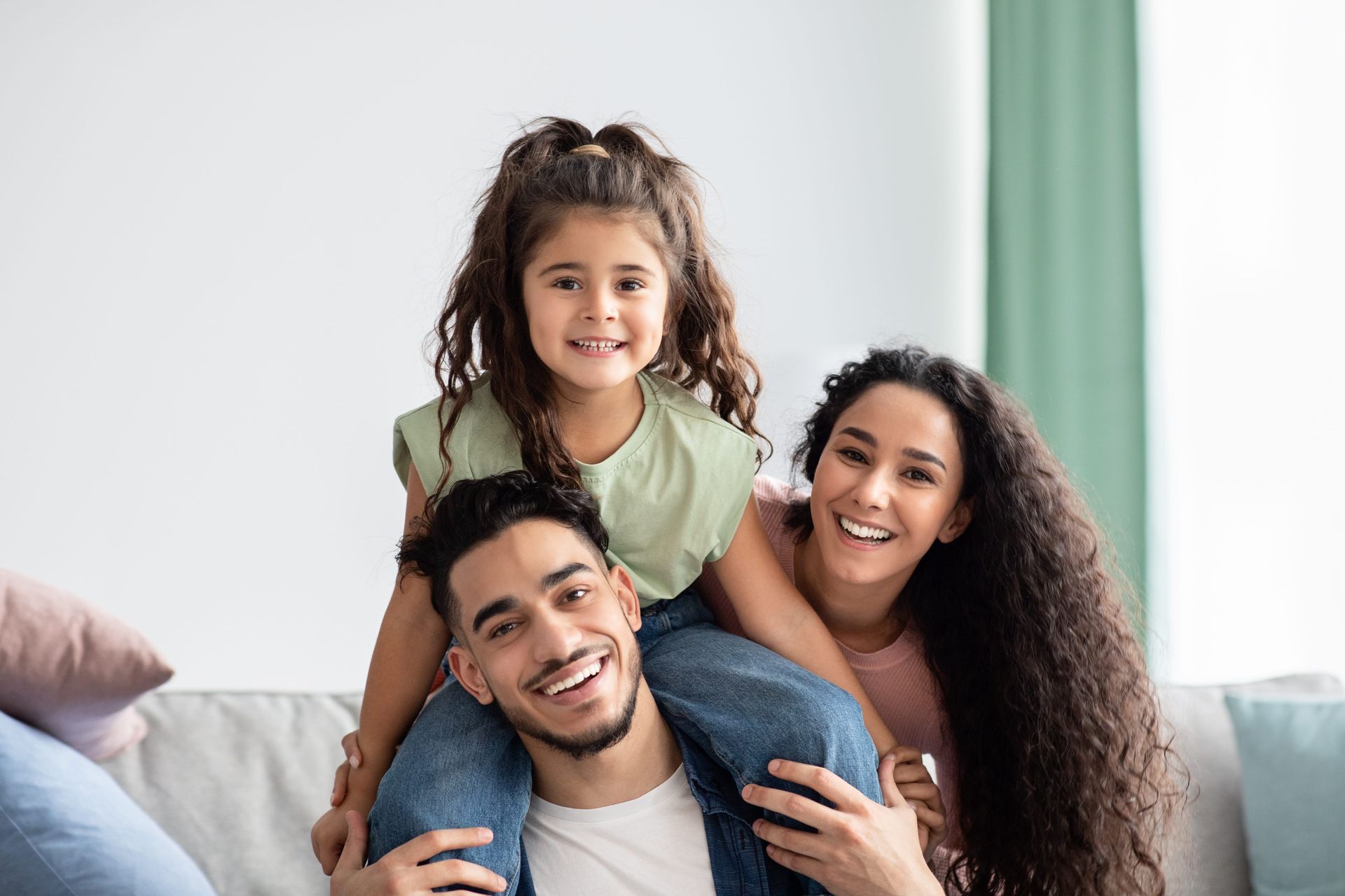 Smiling family sitting on a couch, with a child perched on the adults’ shoulders in a bright living room