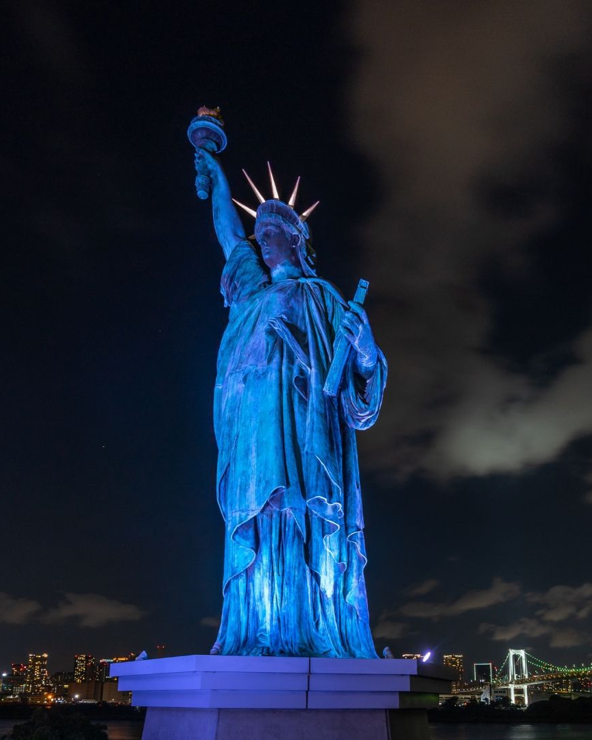 Blue-lit Statue of Liberty at night, standing against a dark cloudy sky with city lights in the distance