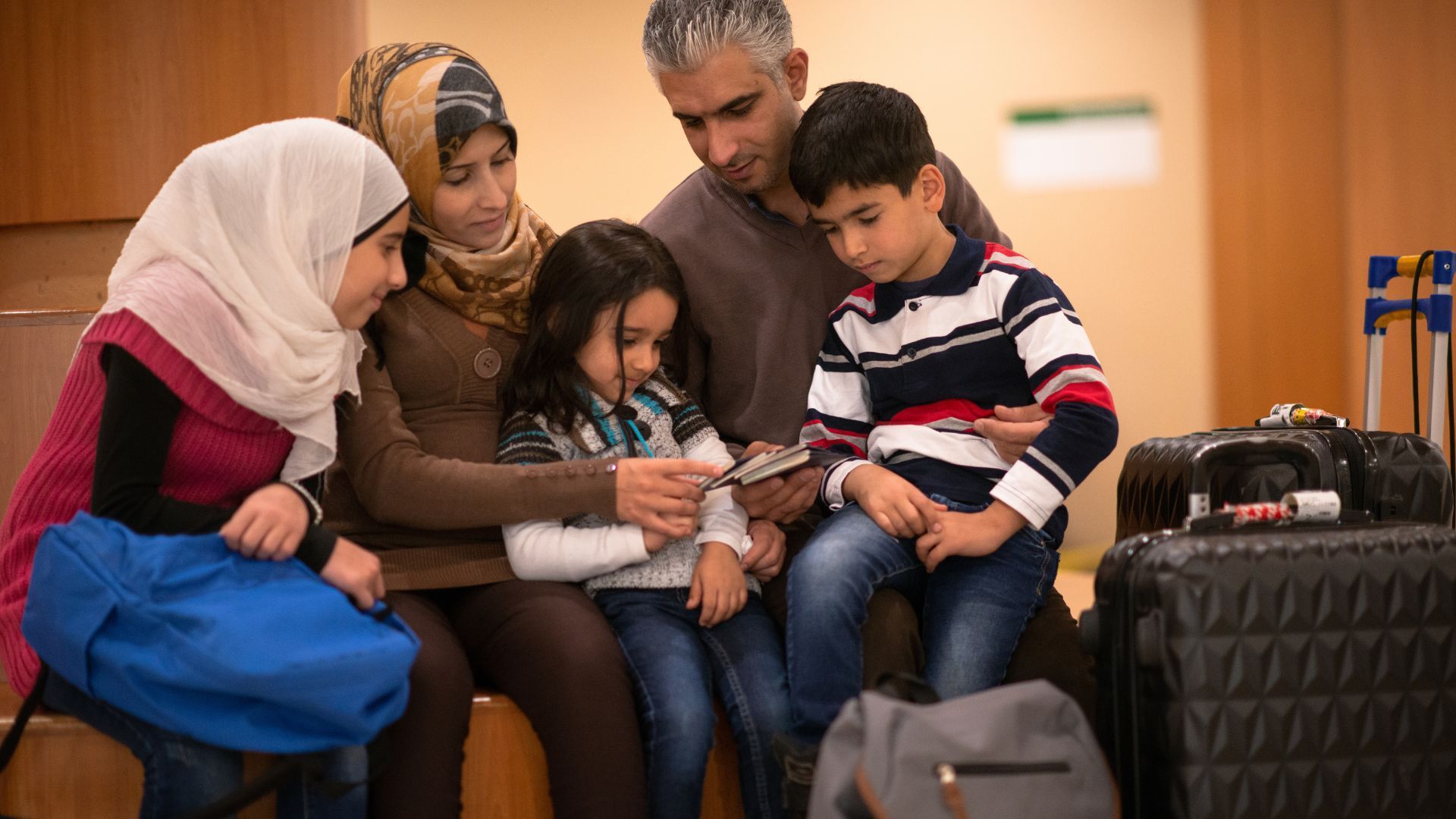 Family gathered around a tablet in an airport lounge, with luggage beside them