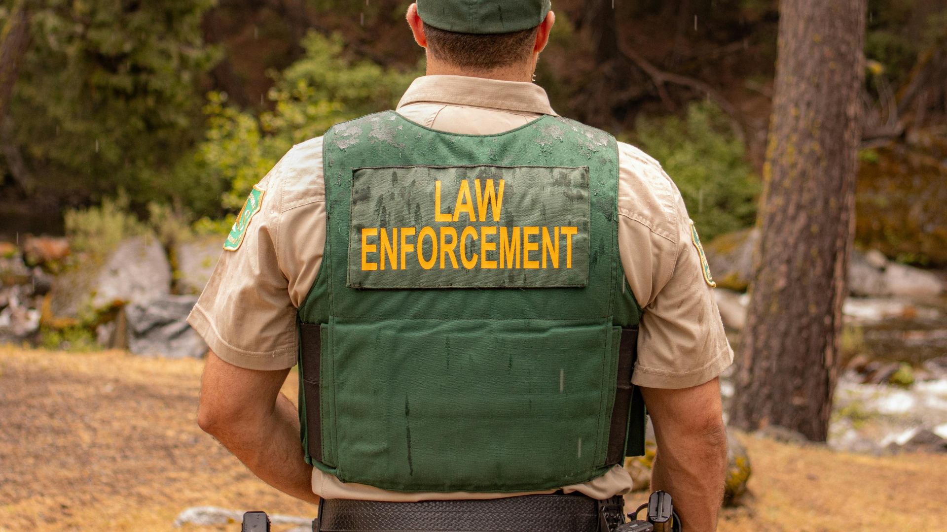Law enforcement officer seen from behind wearing a green vest in a wooded outdoor area