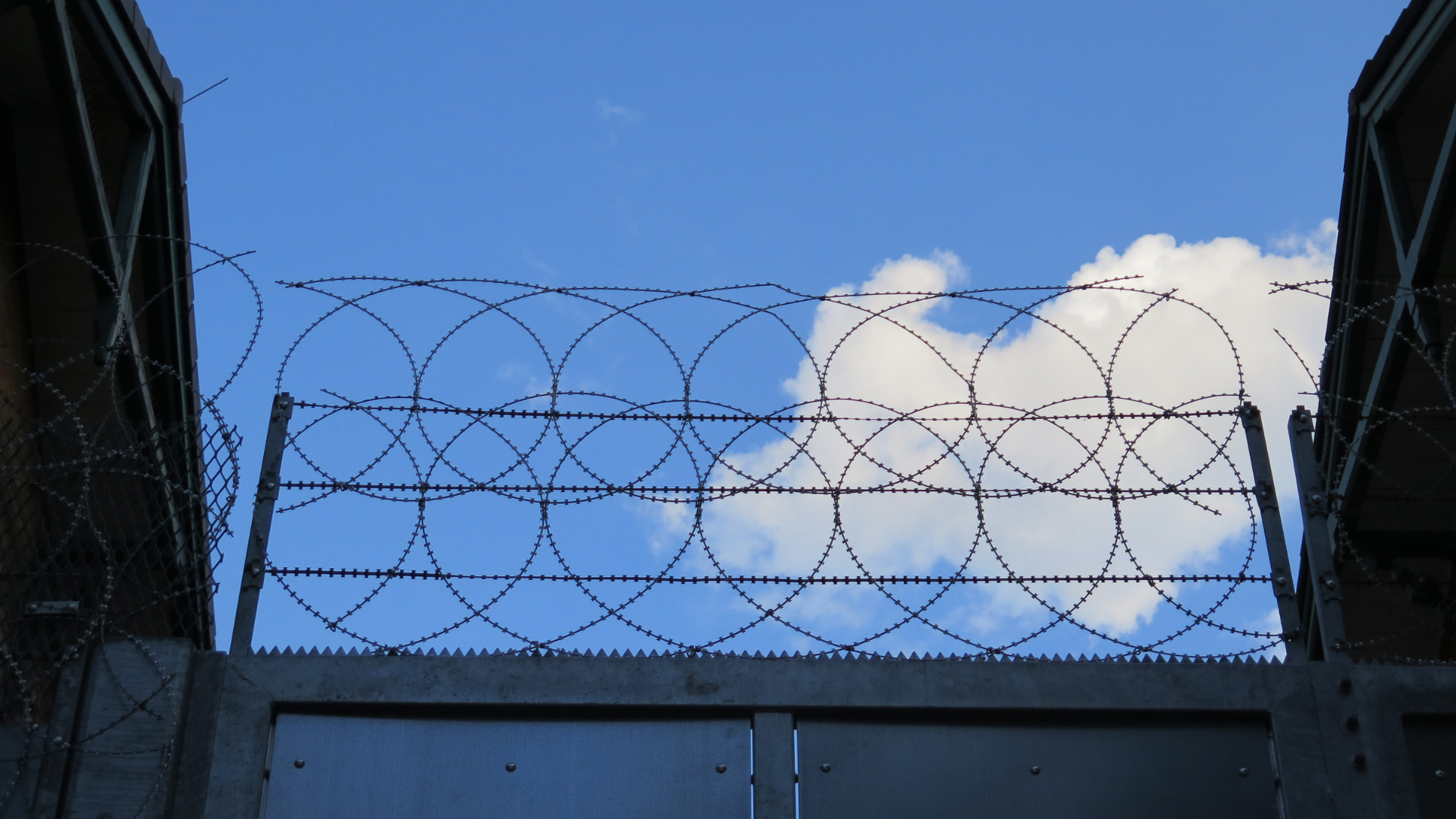 Barbed wire fence silhouetted against a blue sky with white clouds between dark buildings