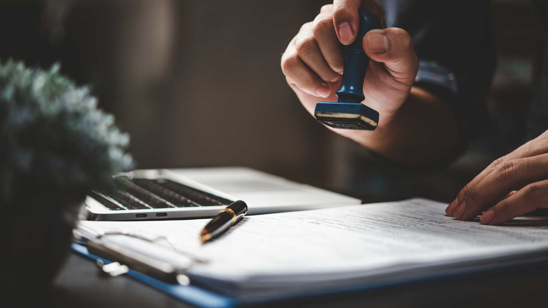 Hand stamping a document beside a laptop and pen on a desk
