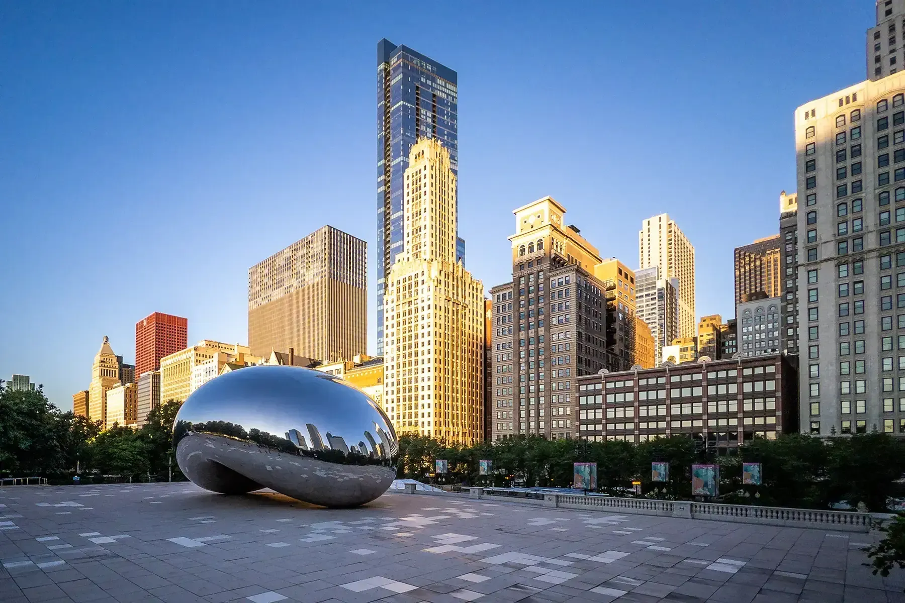 Cloud Gate sculpture in a plaza with sunlit high-rise buildings under a clear blue sky.
