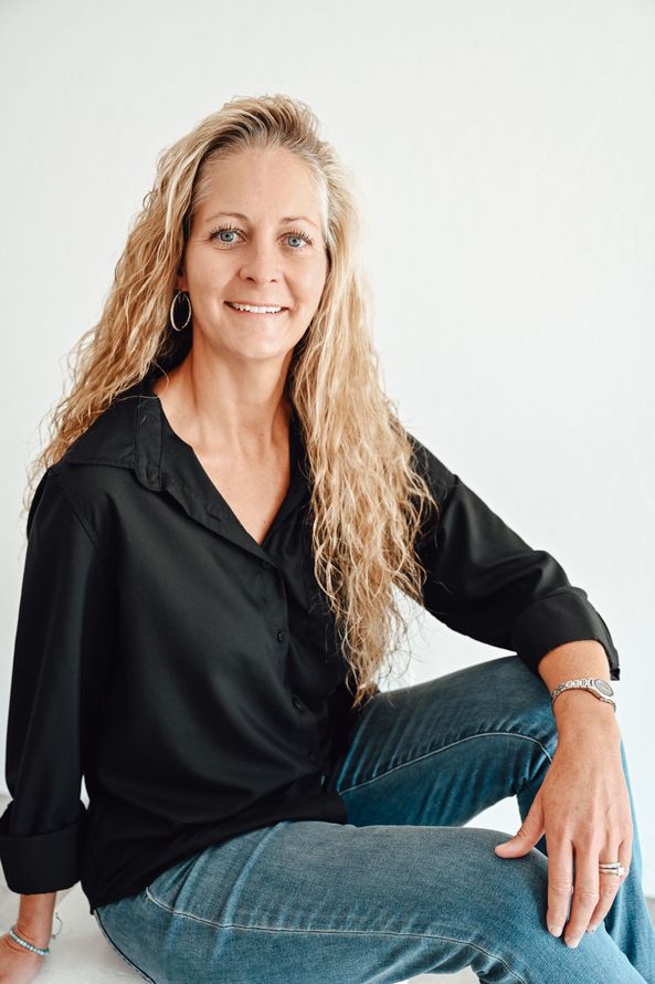Woman seated on the floor in a black blouse and jeans, smiling against a white background