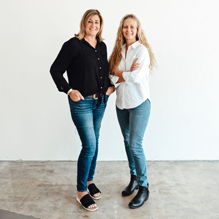 Two women standing together against a white wall, smiling in casual clothes.