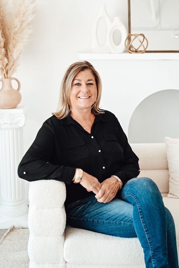 Woman seated on a white chair in a bright living room, wearing a black top and blue jeans.