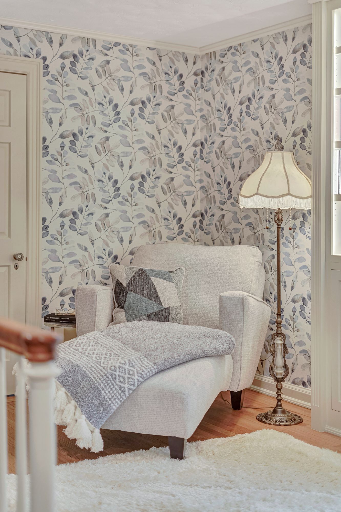 Cozy reading nook with patterned wallpaper, white armchair, blanket, floor lamp, and white rug.