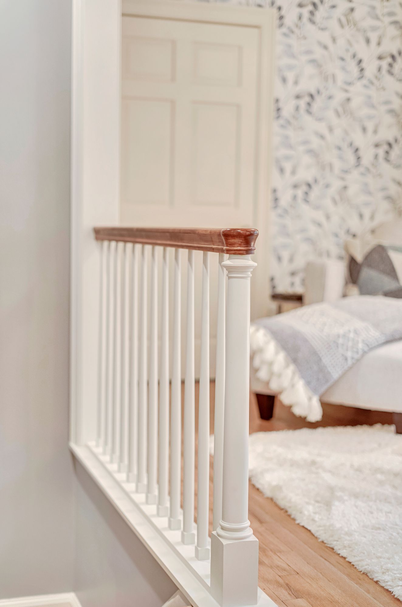 White banister with wooden top in a hallway overlooking a bedroom with patterned wallpaper and a bed.