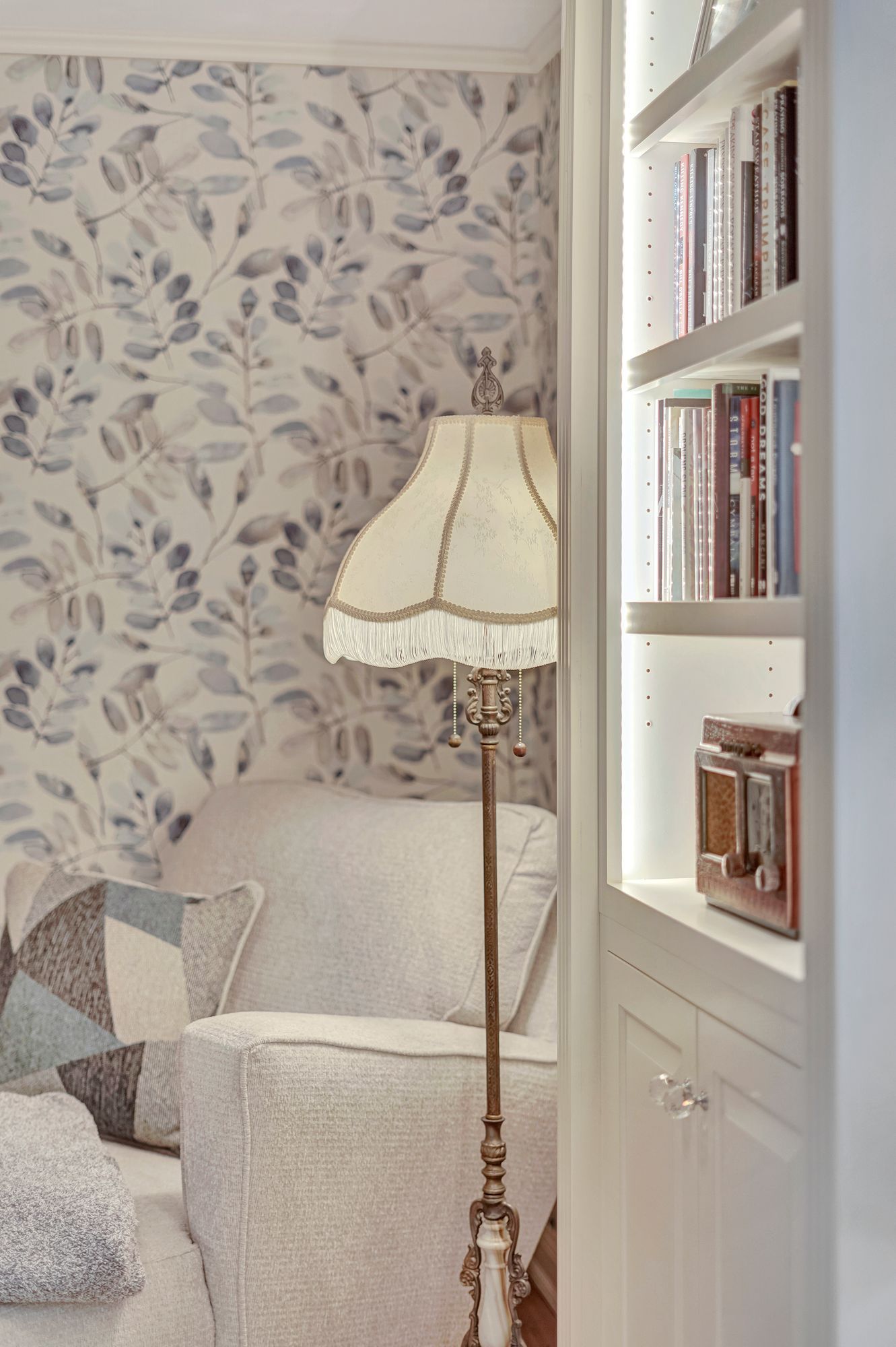 Cozy reading nook with floral wallpaper, a white armchair, and a floor lamp next to a built-in bookshelf.
