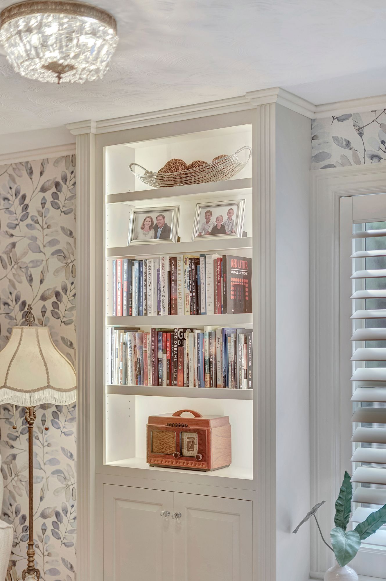 White built-in bookshelf with illuminated shelves displaying DVDs, decor, and a vintage radio. Adjacent to floral wallpaper and a lamp.