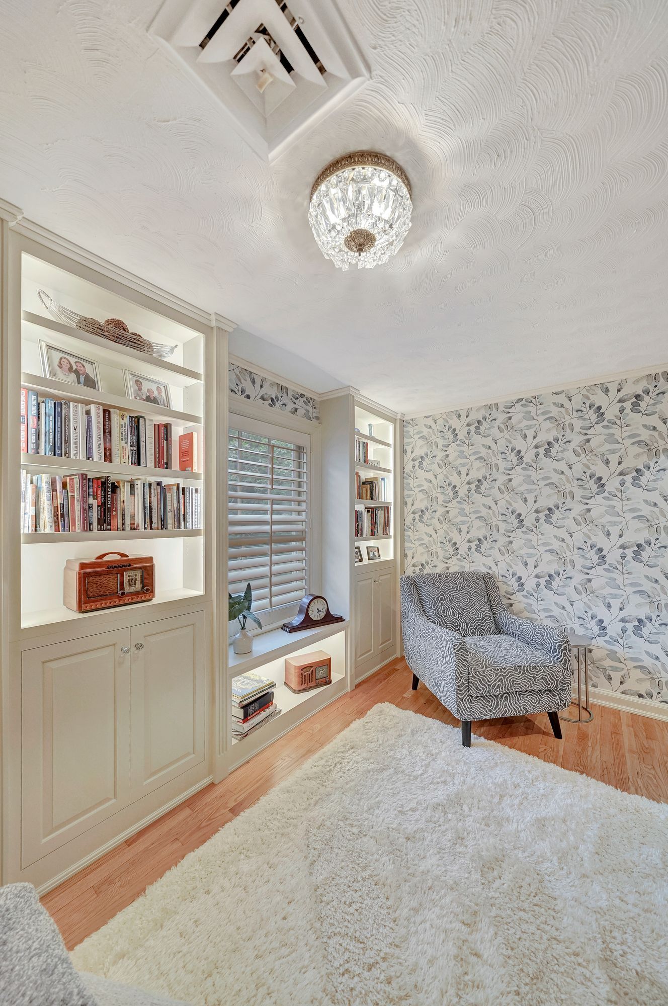 Cozy room with built-in bookshelves, patterned wallpaper, armchair, window with shutters, and fluffy white rug.