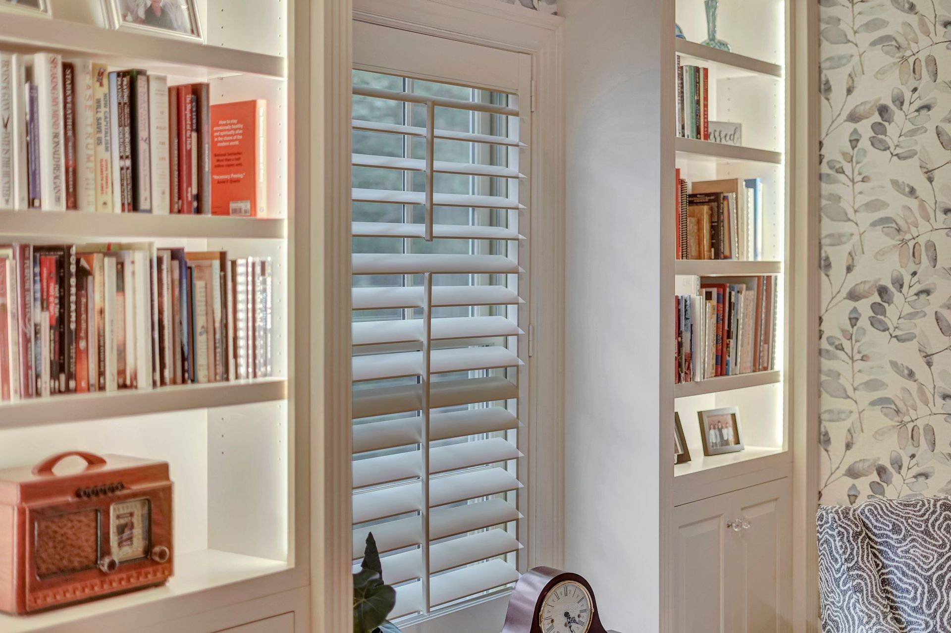 Built-in bookshelves flank a window with white shutters; shelves are lit and filled with books and decor.