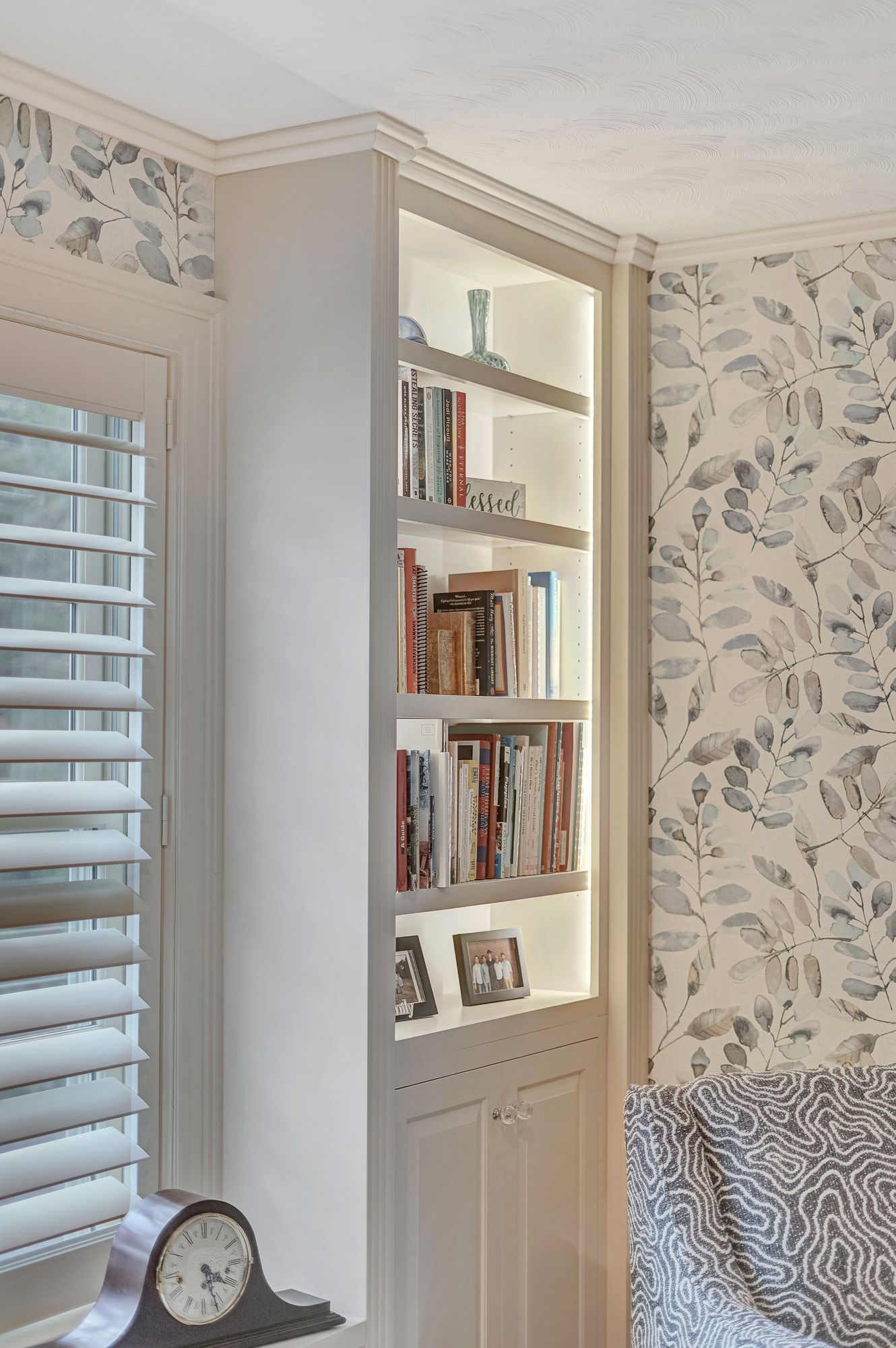 Built-in white bookshelf in a corner with books and a light. Window with blinds and patterned wallpaper.