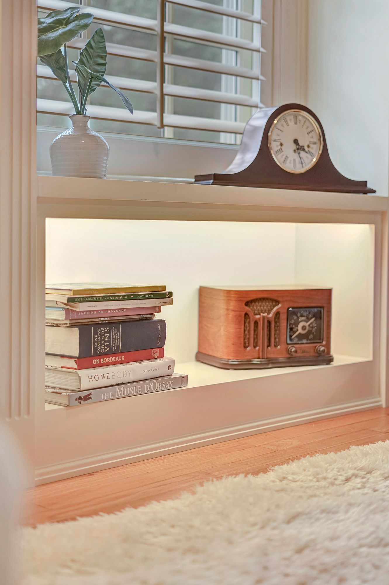 Built-in bookshelf with books, antique clock, and radio; white shelves, soft lighting, and wood flooring.